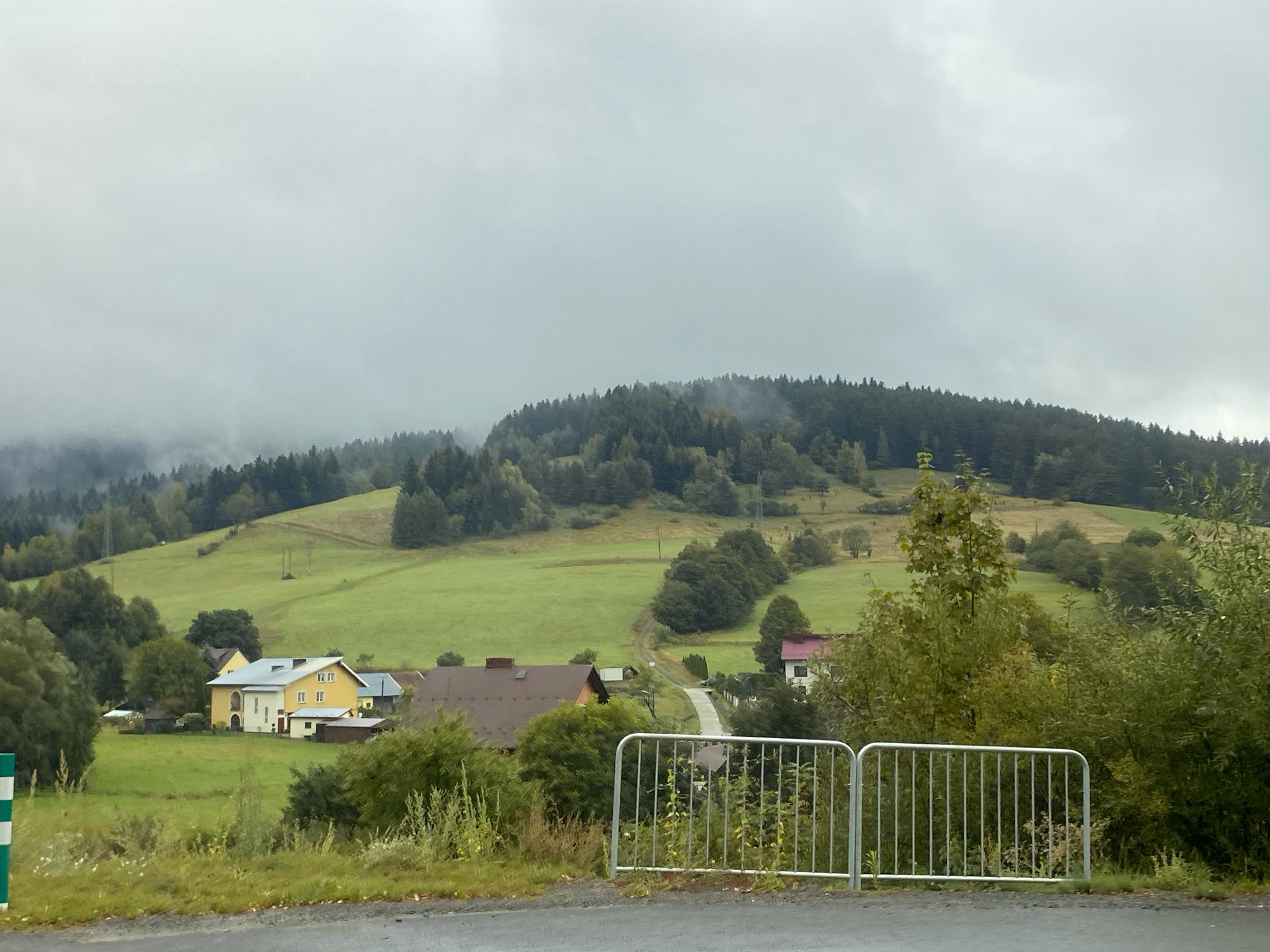 The image depicts a rural landscape in the city of Łabowa, Poland, under an overcast sky. In the foreground, a grey asphalt road is visible, bordered by a green and white striped pole on the left and a silver metal railing on the right. Beyond the railing, overgrown vegetation and grass cover the ground.
The midground features several houses nestled into a gently sloping green hillside. To the left, a bright yellow house with a grey roof stands prominently, accompanied by smaller outbuildings. Further to the right, a house with a dark brown roof is visible, and a lighter colored house with a pink roof is also present. A winding paved road leads through the houses and up the hill.
The background is dominated by a large, tree-covered hill. The trees are dense, with a mix of dark green and lighter foliage. The upper reaches of the hill are shrouded in mist, blending into the grey sky. The overall impression is one of a quiet, rural setting in a hilly region. There are no people visible in the image.