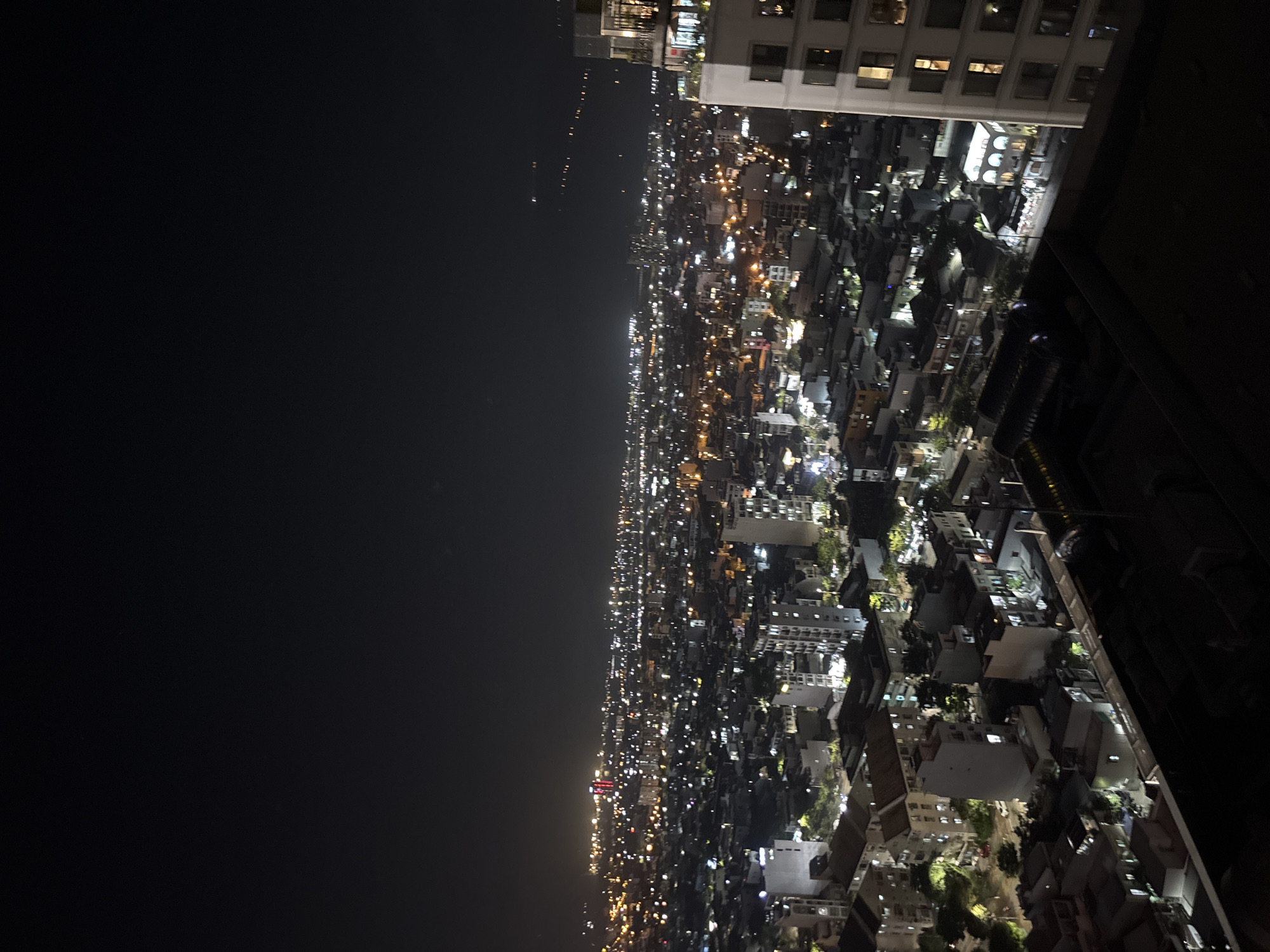 This is a nighttime aerial view of a densely populated urban area, likely in Đà Nẵng, Vietnam, given the provided context of its location. The scene is illuminated by countless artificial lights from buildings, streets, and possibly vehicles. The majority of the structures appear to be multi-story buildings, ranging from smaller residential homes to taller commercial buildings and apartments. The city stretches out into the distance, with lights creating a hazy glow against the dark night sky. Some areas show more distinct clusters of lights, suggesting commercial districts or more concentrated residential zones. The foreground shows the dark rooftops of structures closer to the viewpoint. The overall impression is one of a vibrant, active city at night.