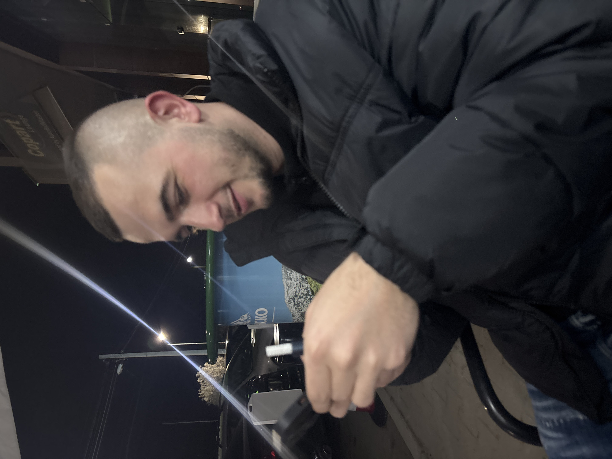 A man is filling up his car at a gas station in Plovdiv, Bulgaria. The man has short hair and is wearing a black jacket. The gas station appears to be in a well-lit area. There is a sign nearby. A black car with a white license plate is being fueled.