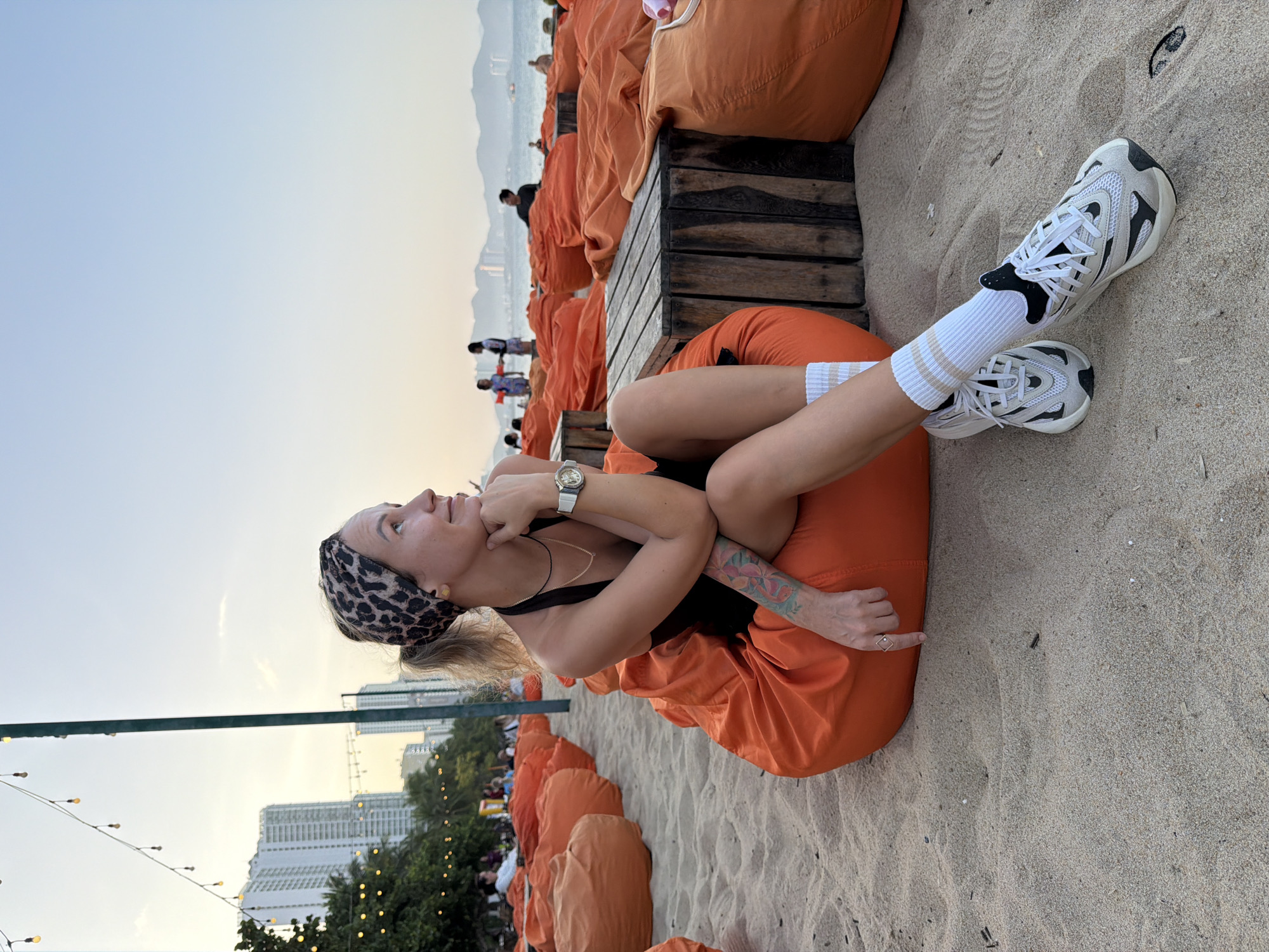 A woman is sitting on a large, orange beanbag chair on a sandy beach in Nha Trang, Vietnam. She is wearing a black dress, white socks with beige stripes, and white athletic shoes. She has a leopard-print headband and a watch on her left wrist. Her right hand is resting on her chin, and she is looking to the right. In the background, there are more orange beanbag chairs, wooden tables, buildings, palm trees, and people in the distance. The sky is a clear blue with a hint of yellow, indicating either sunrise or sunset. The scene appears to be a relaxed, outdoor seating area on the beach.
