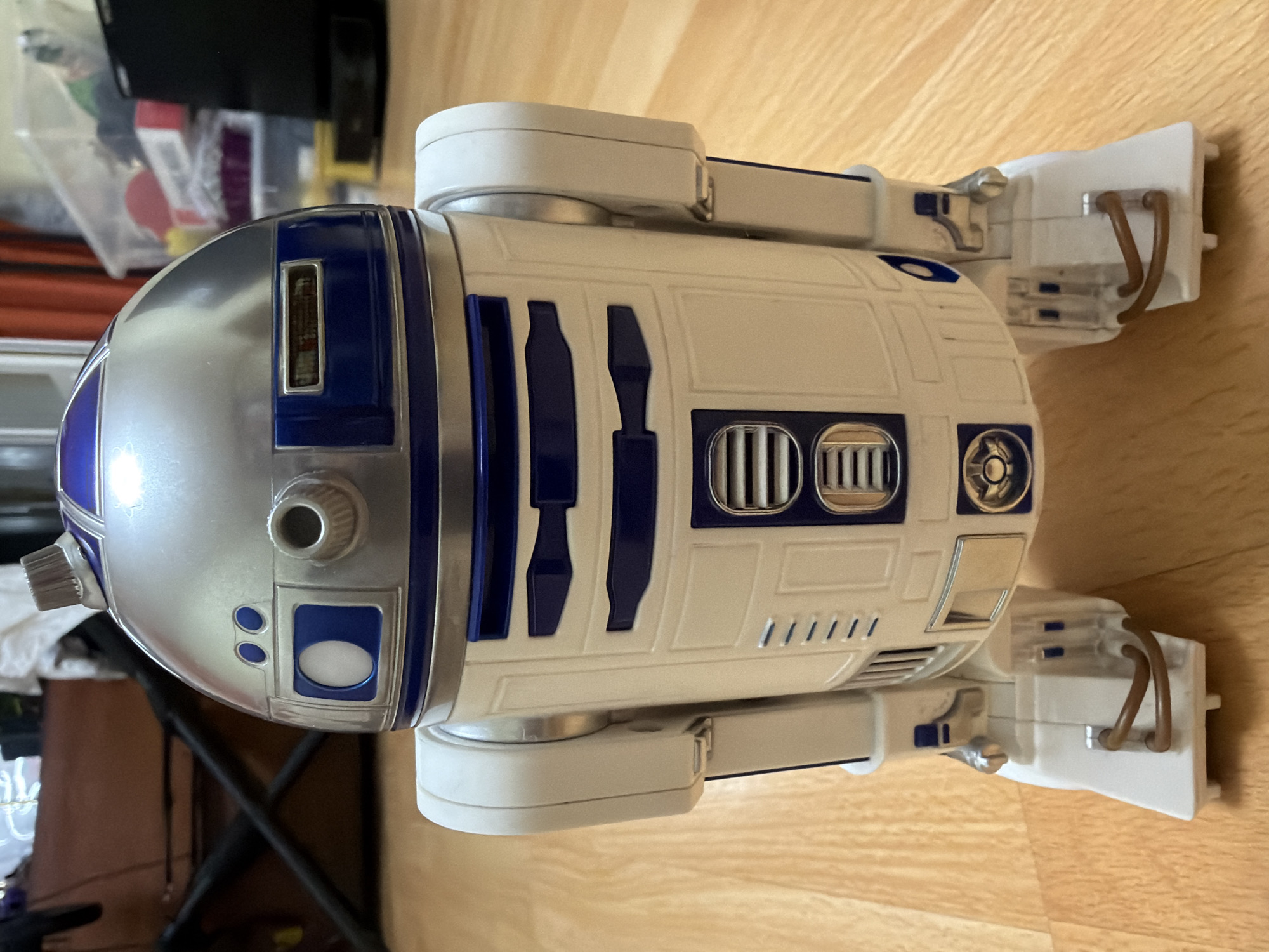 The image shows a close-up, eye-level, front-facing view of an R2-D2 toy figurine standing on a wooden floor. The figurine is white and blue with metallic silver accents on its head and dome. The R2-D2 toy is detailed with various panels, lights, and a dome that swivels. The background is out of focus but appears to be an indoor setting with furniture and storage containers, suggesting a residential or storage area. The wooden floor is visible in the foreground and extends into the background. No people are visible in the image, and no specific location in Kyiv, Ukraine can be determined from the provided visual information.