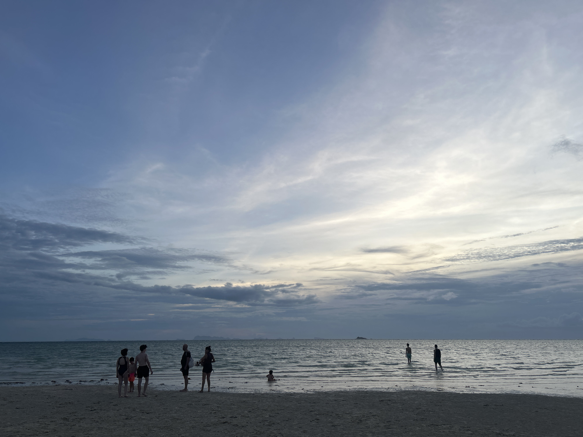 A group of people are gathered on a sandy beach as the sun begins to set over the ocean. The sky is a dramatic canvas of blues and grays, with wispy clouds streaked with golden light. The water is calm, with gentle waves lapping at the shore. In the distance, a few small islands are visible on the horizon. The scene evokes a sense of peacefulness and tranquility, as the day draws to a close.
The group consists of approximately seven individuals, silhouetted against the fading light. They appear to be standing in shallow water or at the water's edge, suggesting a leisurely stroll or a moment of reflection. Their forms are indistinct due to the low light, but their presence adds a human element to the vast seascape. One small figure, possibly a child, is seated in the water further out, seemingly engrossed in their own world.
The overall mood of the image is serene and contemplative. The subdued colors of the sky and sea, combined with the silhouetted figures, create a dreamy and somewhat melancholic atmosphere. The time of day is clearly evening, as indicated by the soft, diffused light and the approaching twilight. The location, a beach in Ko Pha-ngan District, Thailand, further enhances the feeling of a tropical escape and relaxation. There is no visible text in the image.