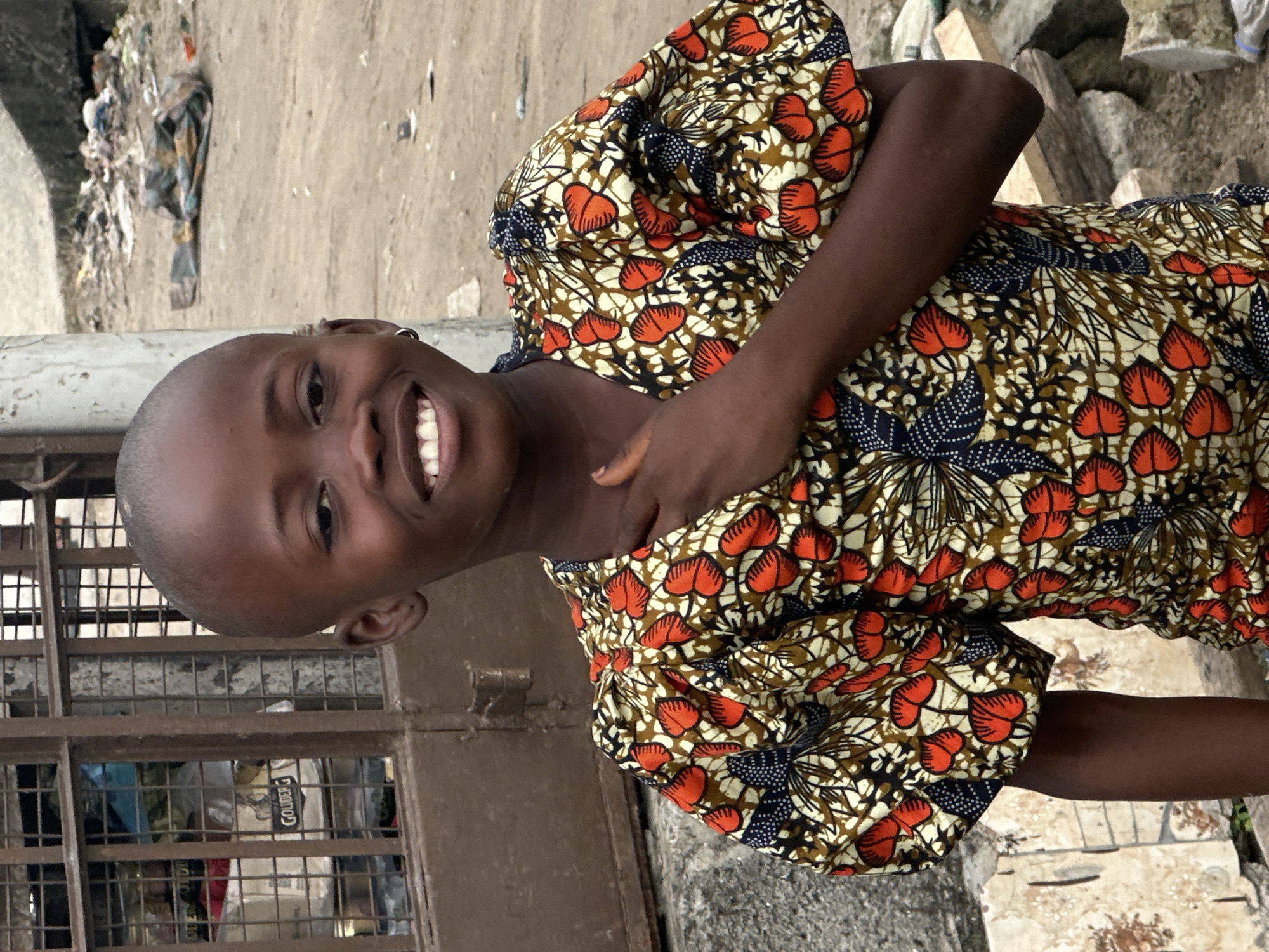 A young person with dark skin and a shaved head smiles broadly, showing their teeth. They are wearing a brightly colored, patterned shirt with puffy sleeves. Their right hand is raised to their chest. The background is blurry and appears to be an outdoor setting with a concrete wall, a metal gate, debris, and a dirt path. The scene is likely in Badagry, Nigeria, based on the provided context.