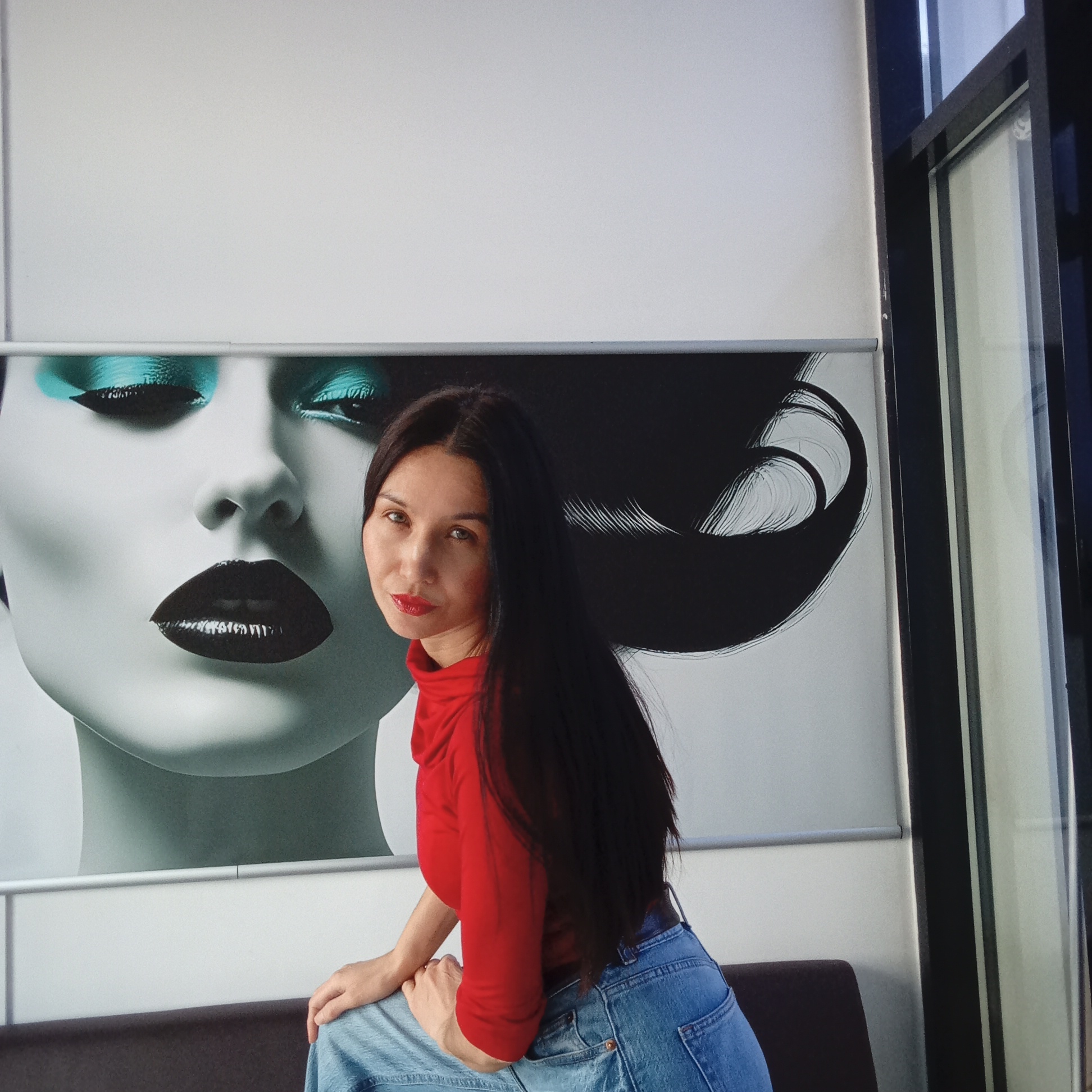 A woman is seated indoors, posing in front of a large, stylized wall poster. She has long, dark hair flowing over her shoulders and is wearing a red long-sleeved top, possibly with a cowl or turtleneck, and light blue denim jeans. Her hands are resting on her lap, and she looks directly at the viewer with a calm, composed expression, her lips painted red.
Behind her, the prominent artwork features a dramatic, close-up depiction of a woman's face. This stylized face is characterized by bright turquoise eyeshadow, glossy black lips, and a striking black hairstyle with a large, swirling design on the right side. The skin tone of the face in the artwork is a pale, cool grey or light blue.
The setting appears to be an indoor commercial or public space, possibly a salon, office waiting area, or retail environment in Izhevsk, Russia. A dark bench or seating area is visible beneath the woman. To the right of the artwork, a dark-framed window or glass partition provides natural light, although the time of day is not explicitly clear. Above the poster is a plain light-colored wall or a rolled-up blind. No text is visible in the image.