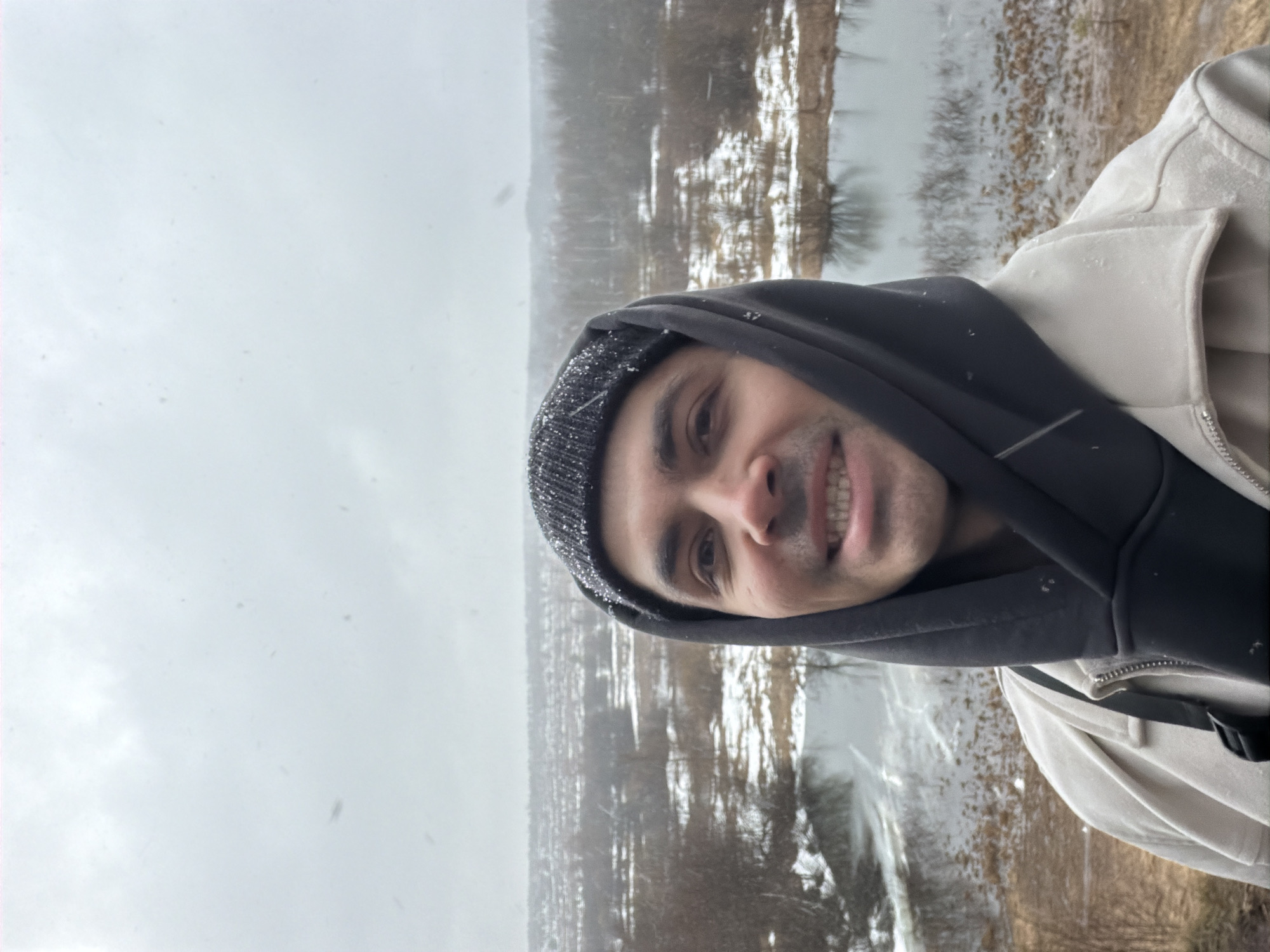 A man is centered in the frame, slightly rotated to the right. He is wearing a black hooded sweatshirt under a light-colored jacket, and a dark beanie with what appears to be snow on it. The man is smiling, revealing his teeth.
The background appears to be a wintery landscape, presumably in Izhevsk, Russia. The area includes what could be a partially frozen river or a snow-covered landscape with trees, visible in the distance. The sky is overcast, and there appears to be light snowfall. The overall setting suggests an outdoor location during winter conditions.