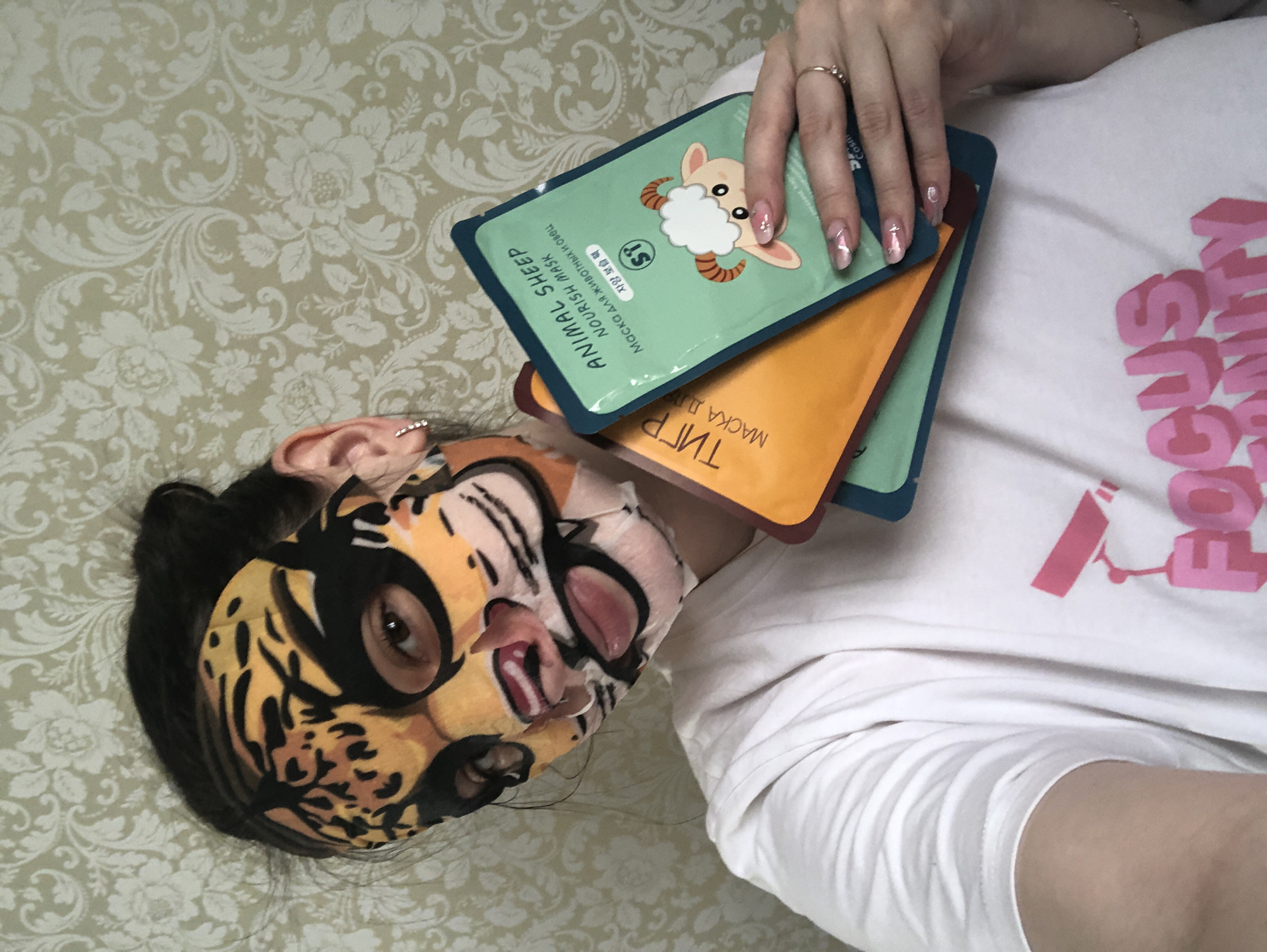 A person is lying down indoors in Noviy, Russia, wearing a tiger-patterned sheet face mask. The mask covers the forehead, cheeks, nose, and chin, leaving the eyes, mouth, and parts of the nose visible. The person has dark hair and is wearing a white t-shirt with partially visible pink text that reads "FOCUS". One hand, adorned with a ring and manicured nails featuring delicate designs, holds three packaged face masks. The top packet is green and features a cartoon sheep, with text "ANIMAL SHEEP NOURISH MASK," "Маска для животных и овец" (Mask for animals and sheep in Russian), and "자양 보습 팩" (Nourishing moisturizing pack in Korean), along with an "ST" logo. Beneath it, an orange packet is partially visible, showing the Cyrillic text "ТИГР" (Tiger) and "МАСКА ДЛЯ" (Mask for). The person's ear is visible, featuring a small studded earring. The background consists of light-colored bedding or wallpaper with an intricate floral and scroll pattern. The image captures a moment of self-care and relaxation.
