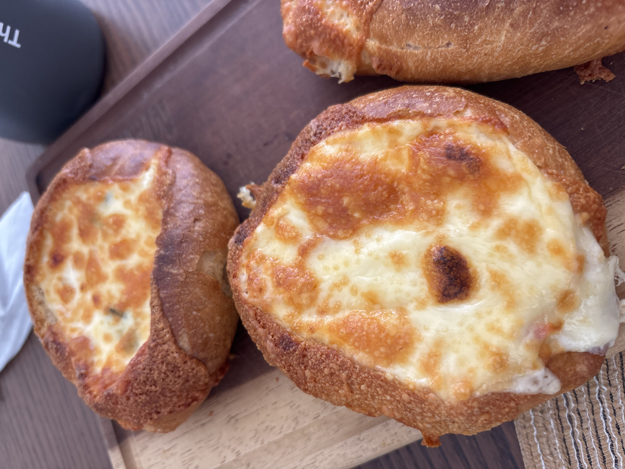 The image displays a close-up, top-down view of several baked goods arranged on a wooden serving tray. Two prominent round, savory pastries dominate the foreground. Each pastry has a golden-brown, crusty bread base generously topped with melted cheese, which shows patches of rich, golden-brown charring from baking or grilling. The pastry on the left has a small visible green inclusion, possibly an herb, beneath its cheese. The larger pastry in the middle foreground features a particularly bubbly and unevenly browned cheese topping. Partially visible at the top of the frame is another elongated baked good, resembling a small baguette, also with a golden-brown crust. A dark-colored mug with inverted white text, showing characters like "TH" or "HT," is visible in the upper left corner. The scene suggests an indoor setting, possibly a cafe or a home, presenting these items for consumption.