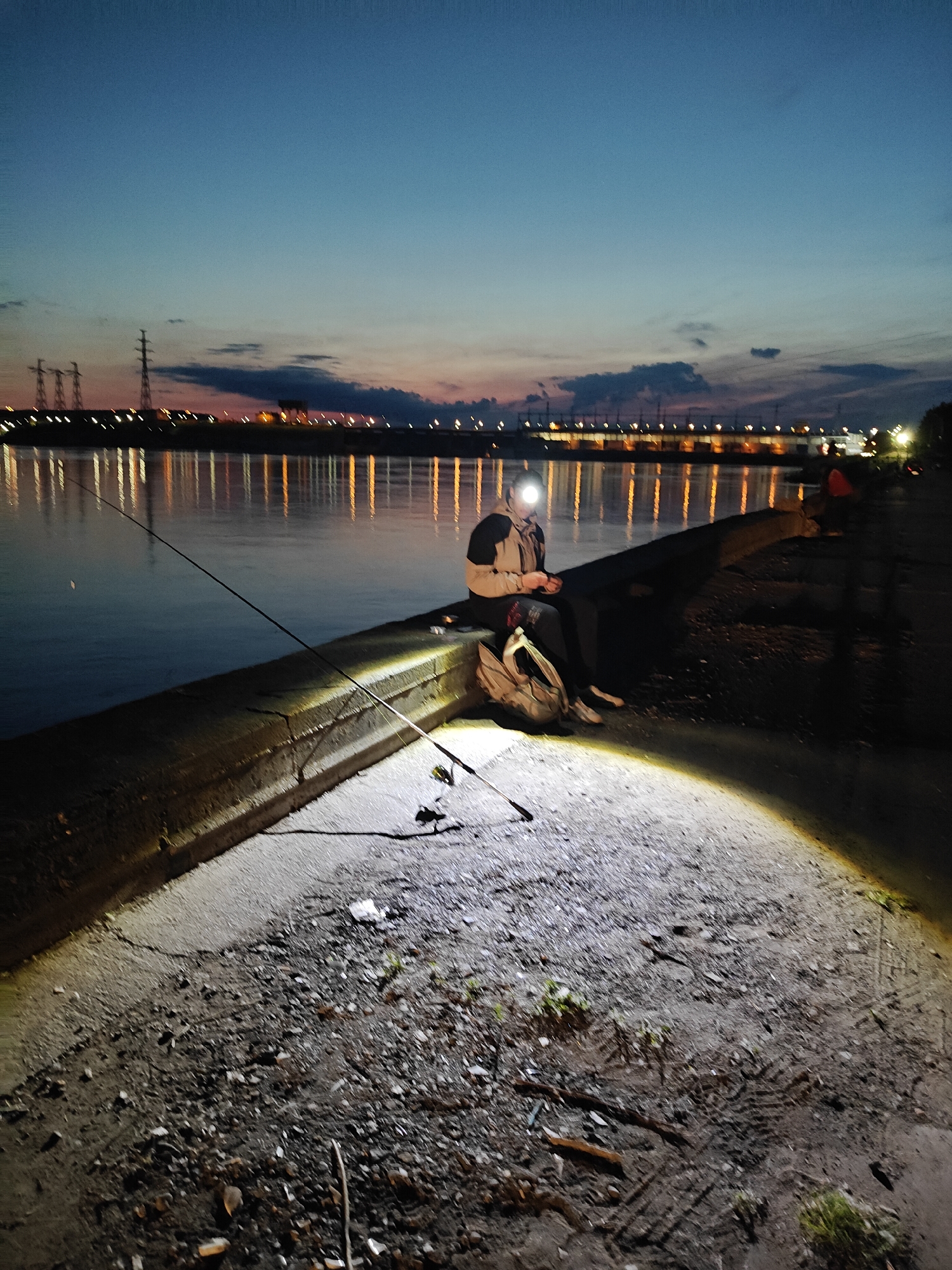 This image captures a nighttime scene of a person fishing by a body of water in Chaikovsky, Russia. The time of day is late evening or early night, characterized by a dark blue sky that transitions to a faint orange and pink hue at the horizon, suggesting the last remnants of twilight.
The primary subject is a lone individual seated on a concrete embankment or pier on the left side of the frame. They are wearing a jacket and a headlamp, which emits a bright, conical beam of light onto the rough, gravelly ground in front of them. A fishing rod is propped up, extending into the calm water. A backpack rests beside the person on the embankment.
Across the body of water, possibly a river or reservoir, numerous artificial lights from buildings or industrial facilities twinkle on the distant shore, casting long, vertical reflections that dance on the water's surface. Silhouettes of power lines and transmission towers are visible against the twilight sky. The immediate foreground consists of a coarse, uneven path or ground composed of gravel, dirt, and some small debris, strongly illuminated by the headlamp. The overall atmosphere is quiet and focused, typical of night fishing. No visible text is present in the image.