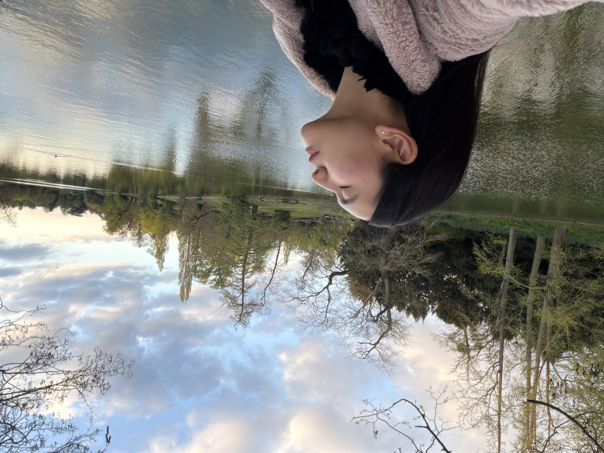 A rotated outdoor shot depicts a woman in a pink jacket with black lining, positioned in front of a lake and trees. The location is likely the Bois de Vincennes in Paris, France. The sky is visible with clouds.