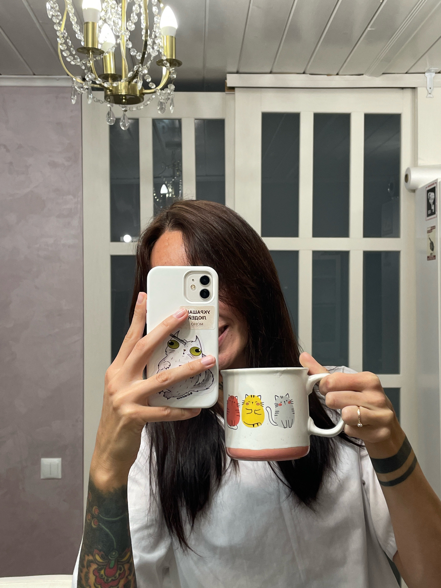 A woman is taking a selfie in a room with a chandelier, white sliding doors, and a purple wall. She is holding a white iPhone in her right hand and a white mug with cat illustrations in her left hand. She is wearing a white t-shirt and has tattoos on her left arm and a black band tattoo on her right forearm. The mug has three cats: one red, one yellow, and one gray. The phone case has a sticker of a white cat with green eyes.