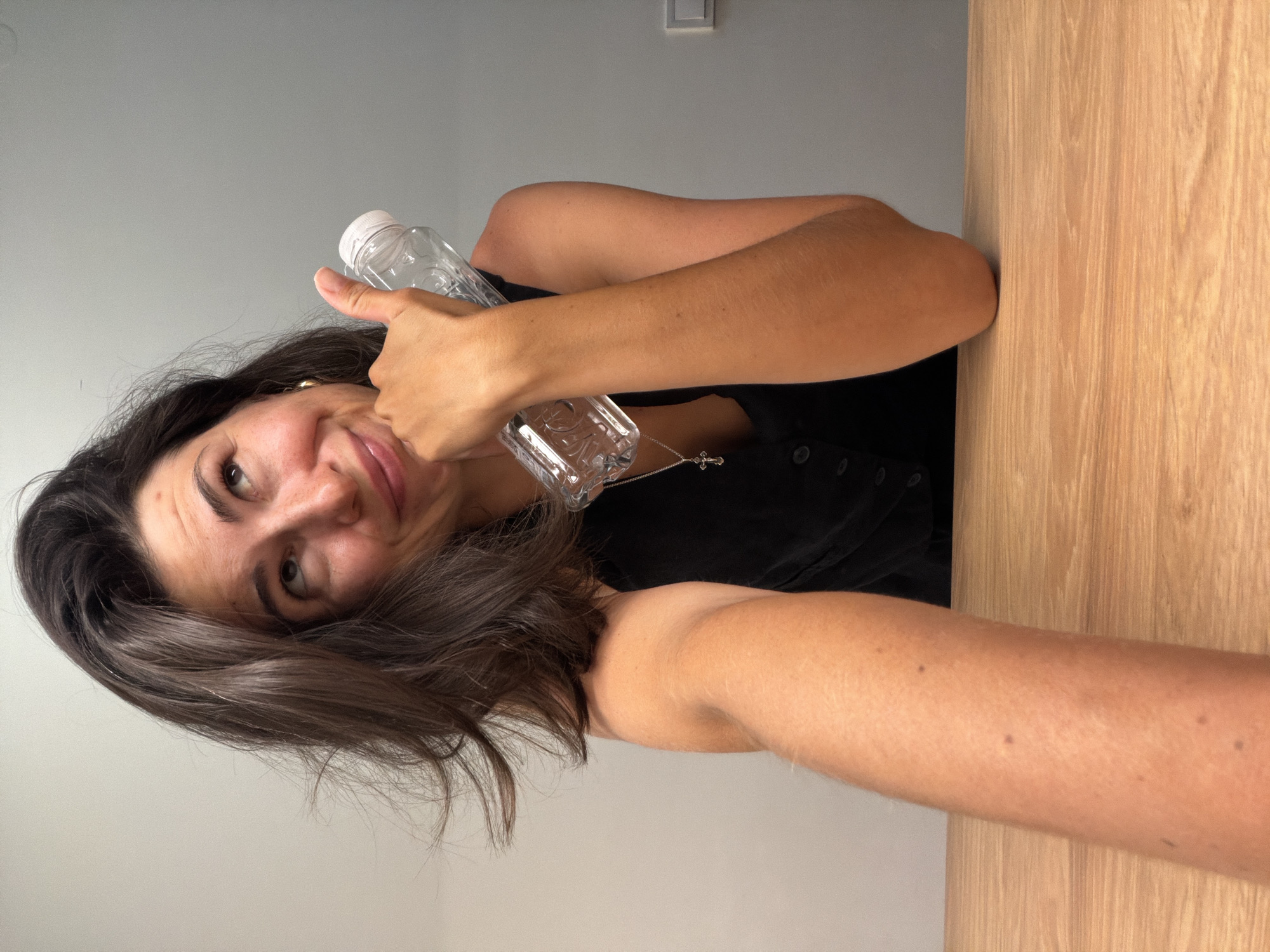 An indoor, selfie-style image captures a woman with dark brown hair and a tanned complexion, situated in Budva, Montenegro. She looks up towards the camera with a subtle, friendly smile, her head tilted. She wears a dark top and a silver cross necklace. Her right hand is raised, giving a thumbs-up gesture, while her left hand holds a clear plastic water bottle with a white cap, resting against her chest. The background consists of a plain light gray wall on the left and upper sections, where a white light switch is visible. To the right, a light-colored wooden surface, possibly a table or door, forms the backdrop. The scene is brightly and evenly lit. No visible text is present in the image.