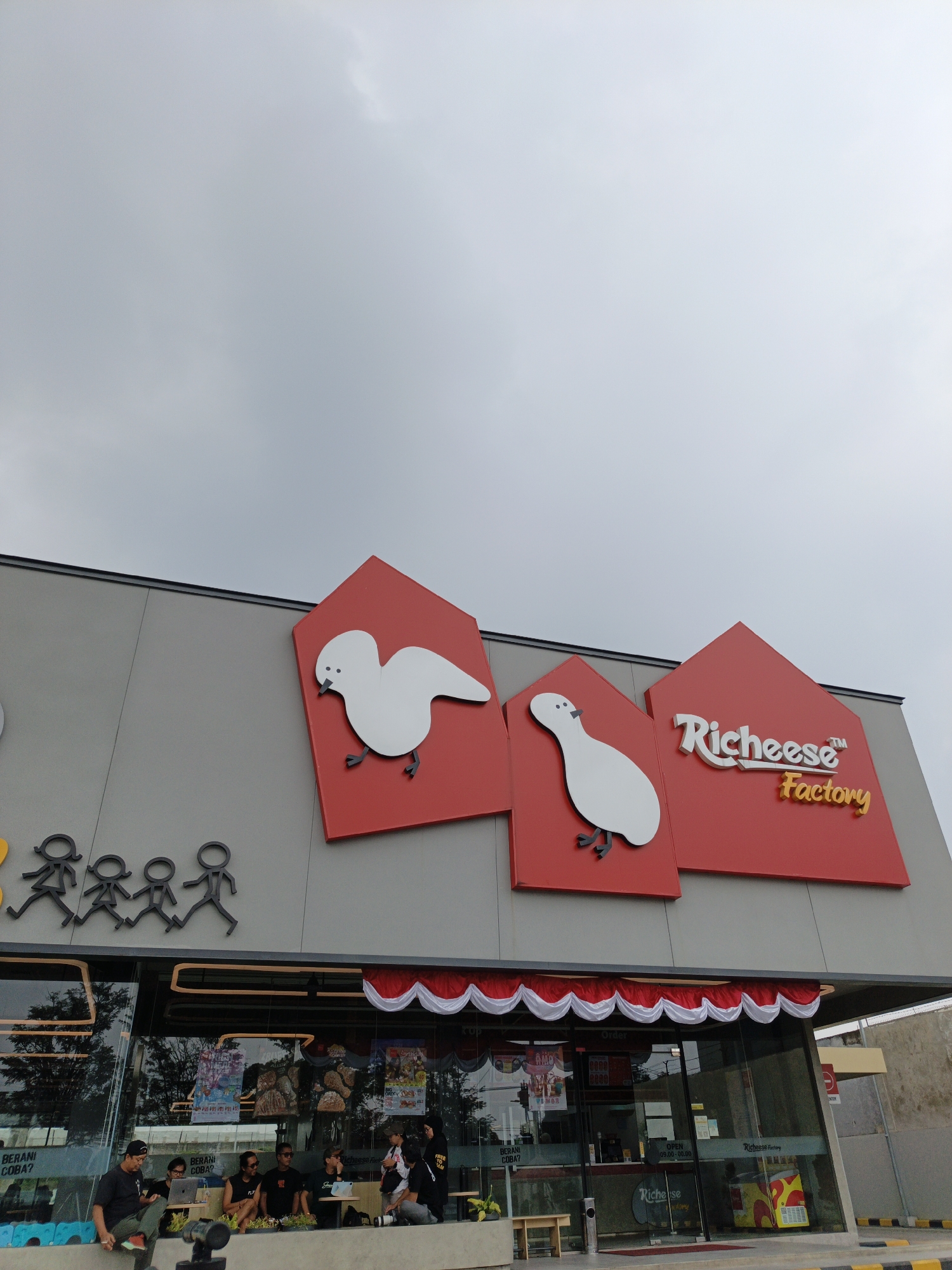 The image shows the exterior of a Richeese Factory restaurant in Lembang, Indonesia. The building has a modern facade with a gray wall and large glass windows. Prominently displayed on the upper part of the building are three red, house-shaped signs, two of which feature stylized white birds. The middle sign has the "Richeese Factory" logo in white and yellow text. Below the signs, there is a row of black stick figures representing a family.
In front of the building, several people are gathered, some seated on a low concrete wall. They appear to be a group of friends or acquaintances. One person is using a laptop, and another is holding a camera. They are casually dressed, suggesting a relaxed atmosphere. Inside the restaurant, through the glass windows, various posters and displays are visible, along with the restaurant's interior decor. The overall environment suggests a public space, likely a tourist or recreational area given the location in Lembang. The sky is overcast, indicating potentially cloudy weather.