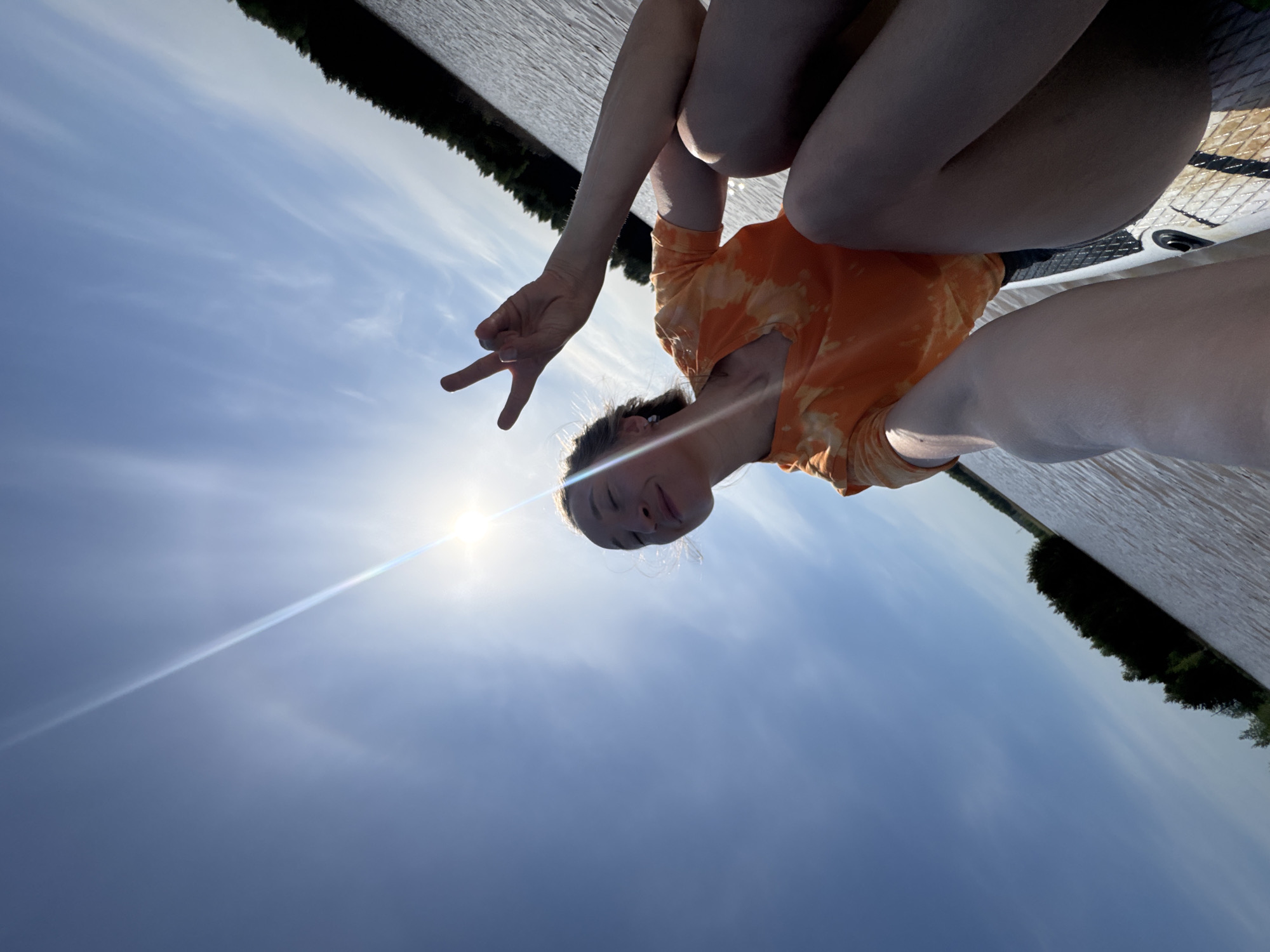 A young person is taking a selfie outdoors on the water. They are sitting on a paddle board and making a peace sign with their hand. The person is wearing an orange tie-dye shirt. The sky is bright blue, and the sun is shining. In the background, there is water and trees. The image is angled upward, and the scene is rotated in the frame. There is no mention of Novyi, Russia in the image.