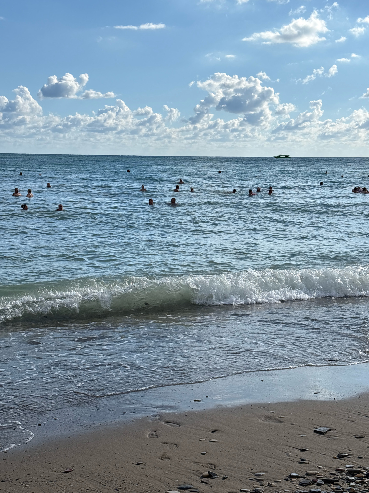 The image depicts a crowded beach scene in Arkhipo-Osipovka, Russia, under a partly cloudy blue sky. The sea is calm with small waves breaking on the shore. Numerous people are visible in the water, engaged in swimming and recreational activities. A small boat is positioned on the horizon. The beach itself is composed of sand and pebbles, with footprints evident in the wet sand near the water's edge. The overall atmosphere suggests a warm and sunny day at a popular seaside resort.