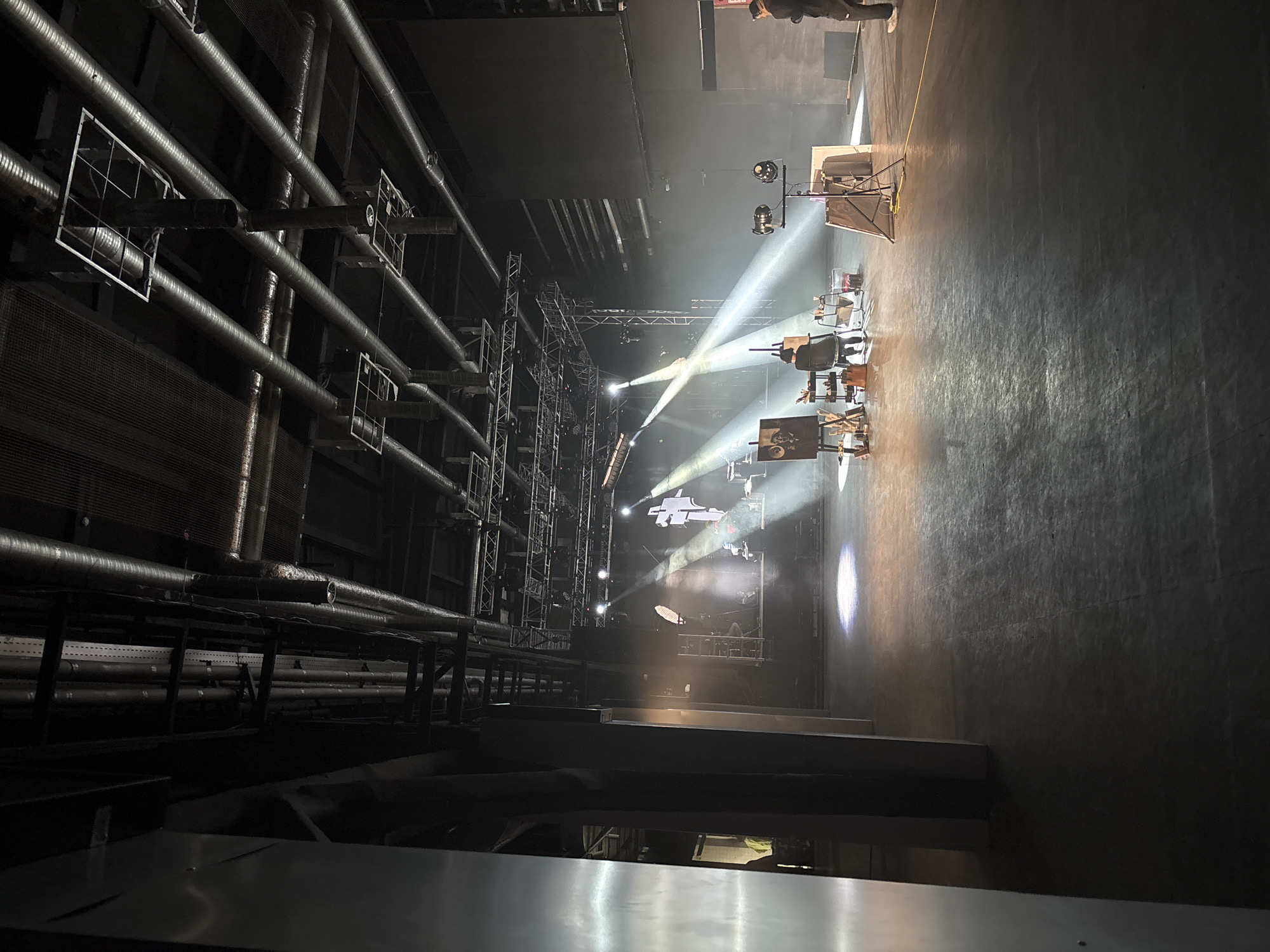 The image displays a vast, dimly lit indoor performance venue, likely a theater or concert hall, captured from an elevated perspective looking down onto the stage.
On the left side of the frame, several large, dark grey cylindrical pipes run horizontally along a wall, with white, square-gridded frames attached to some. Further into the distance on the left, intricate metal truss systems are visible, indicative of stage rigging for lighting and set elements.
The central and right areas are dominated by a stage setup. Bright spotlights pierce through atmospheric haze or smoke, projecting strong, conical beams of light onto the performance space. The stage floor is dark and subtly reflective. Several dark chairs are arranged on the stage, suggesting seating for a musical ensemble or performers. Towards the front of the stage, two easels stand, with one clearly displaying a black and white portrait of a man, possibly with dark hair and a beard, wearing a suit. Other stage equipment, such as monitors or speakers, are discernible in the background of the stage.
The scene depicts a professional environment for a performance, rehearsal, or sound check, characterized by dramatic lighting. A person stands near the stage at the far upper right, and another person's lower legs are visible closer to the camera. No discernible text is visible within the image. The location context is Moscow, Russia.