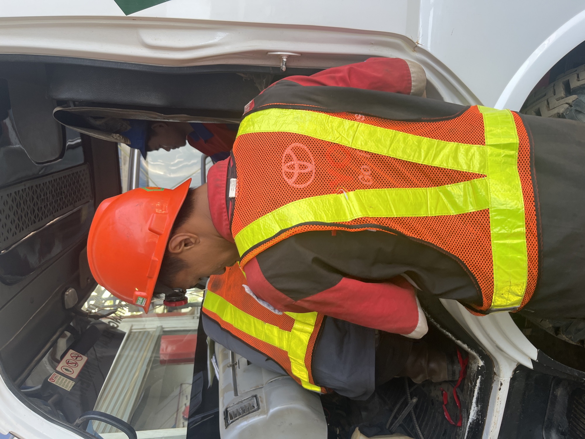 Two individuals are inside the cabin of a white truck. The person in the foreground is wearing a red shirt, a black vest with orange reflective stripes, and an orange hard hat. They are facing away from the camera and appear to be working on the truck's interior. A partial Toyota logo is visible on their vest. The second individual, partially visible in the background, is wearing a blue cap and a red shirt. The truck's interior is cluttered, with some debris on the floor. Outside the truck, a section of a gray building with horizontal metal siding is visible, suggesting an industrial or construction setting. The scene is outdoors, and the lighting indicates it is daytime.