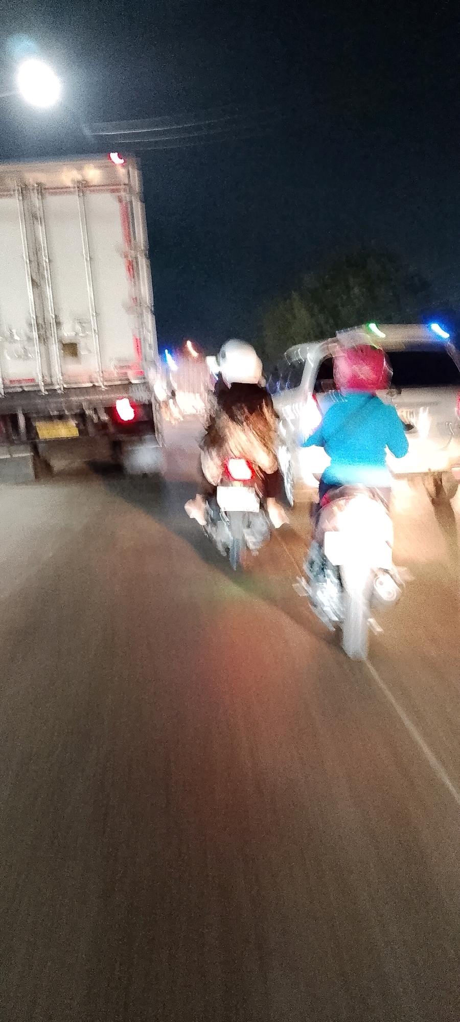 An out-of-focus, nighttime photograph captures traffic on a road in Cileungsi, Indonesia. Two individuals are riding motorcycles in tandem, one in front and one behind, on a dark asphalt road. The lead motorcycle rider is wearing a white helmet and a dark jacket, while the rider behind them wears a bright blue jacket and a red helmet. Both motorcycles have their brake lights illuminated. To the left of the motorcycles is the rear of a large, white semi-trailer truck. The truck has red taillights and some reflective white and red striping. To the right of the motorcycles, a white vehicle, possibly an SUV, is visible with illuminated blue and green lights on its roof. The sky is dark, suggesting nighttime, and a bright light source, possibly a streetlamp, is visible in the upper left corner of the frame. Trees are faintly visible in the background. The image's blurriness suggests motion, either from the camera or the vehicles.