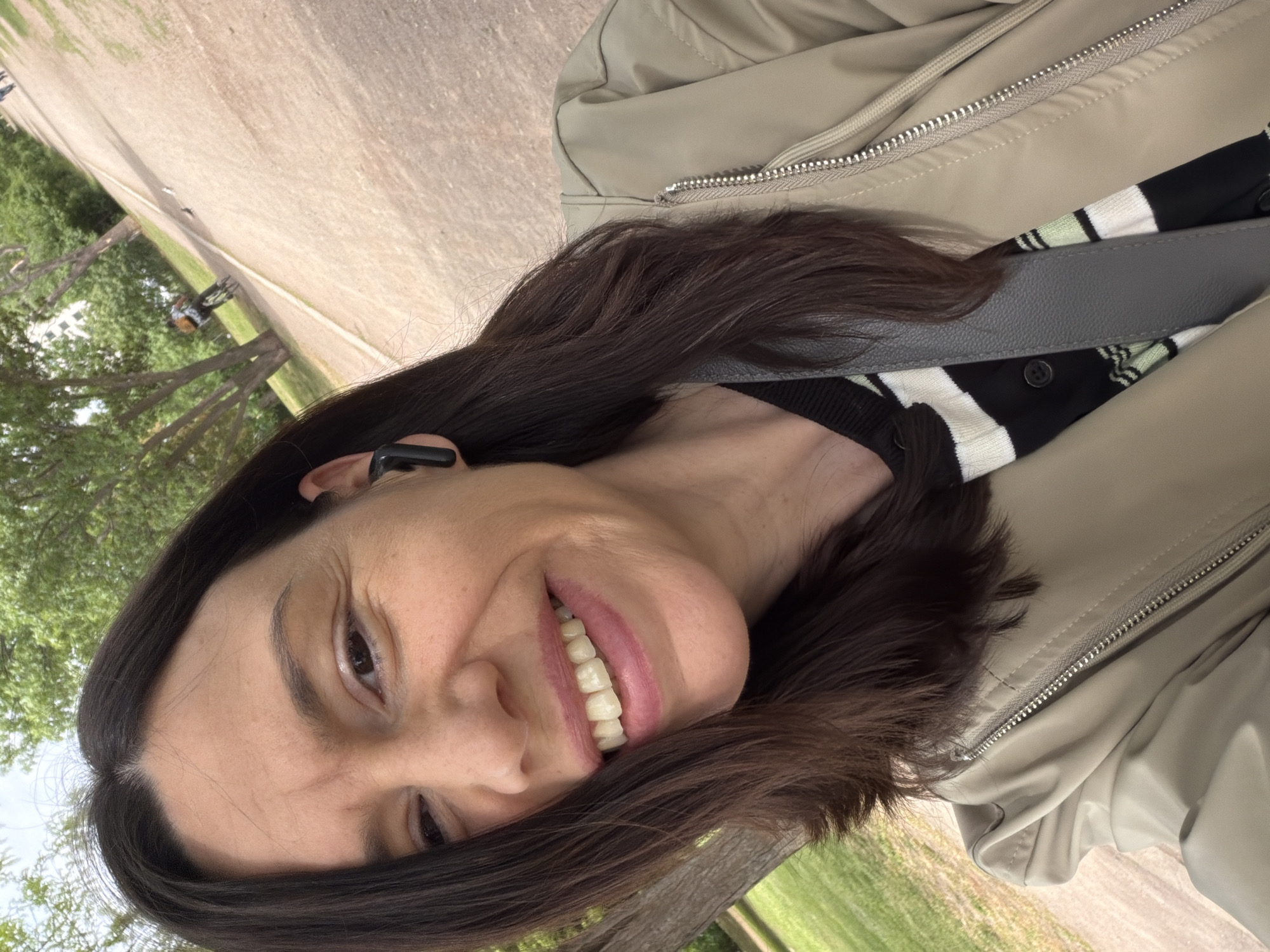 A woman is seen in a close-up, slightly angled selfie. She has dark brown hair, fair skin, and is smiling. She is wearing a black and white striped shirt, a light beige jacket with a visible zipper, and what appears to be a grey leather bag strap across her chest. An earbud is visible in her left ear. The background shows a street or path with green vegetation alongside it, suggesting an outdoor environment. There is no specific information that identifies the location as Sofia, Bulgaria.