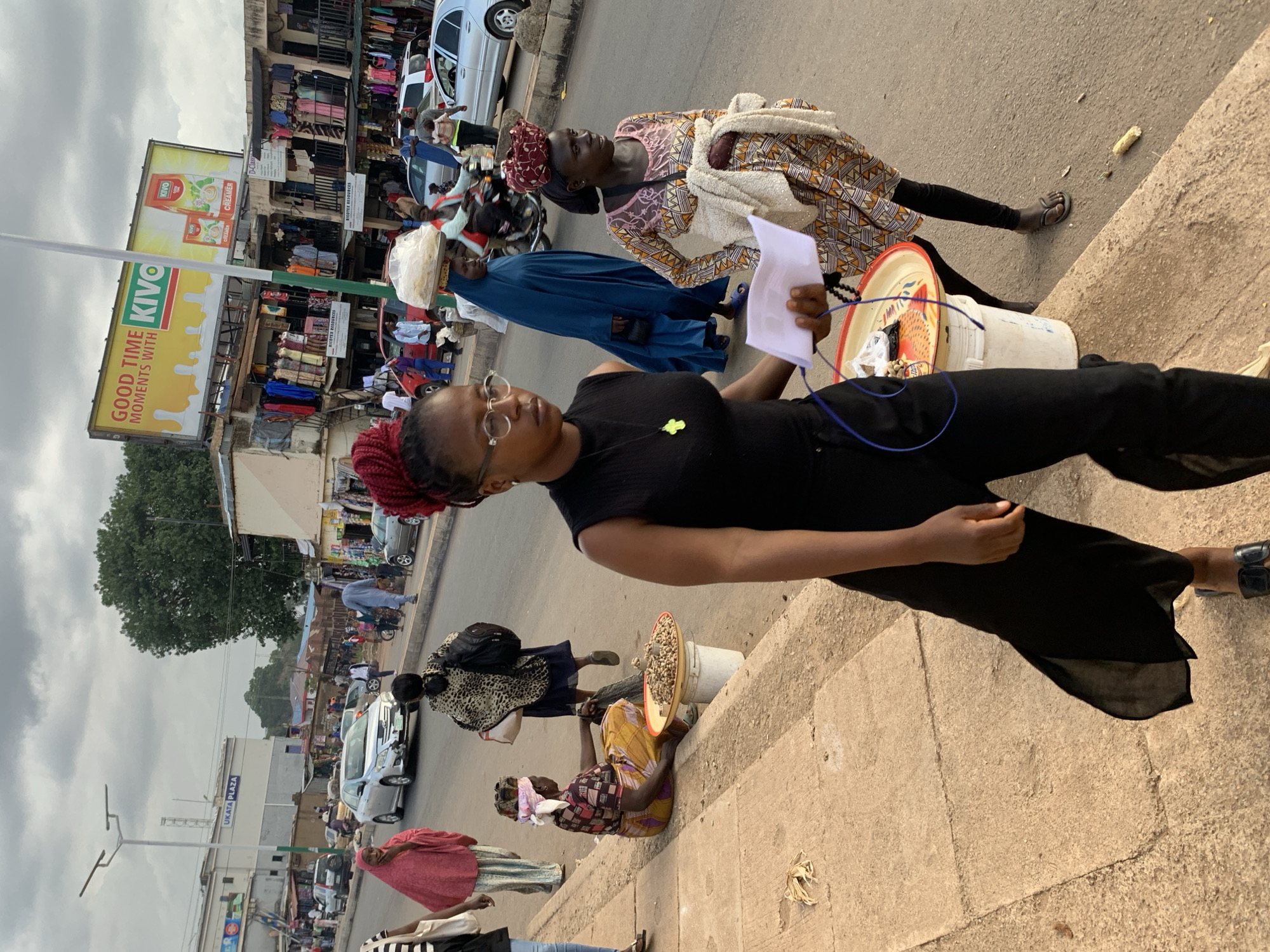The image depicts a street scene in Jos, Nigeria. A woman in a black outfit and glasses is the primary subject, holding a piece of paper and looking forward. She appears to be standing on a concrete sidewalk. Behind her, two other women are visible. One, wearing a blue garment and a headscarf, is carrying a tray on her head, possibly with bread. The other woman, in a patterned dress and headscarf, holds a bundle of cloth. In the background, a busy marketplace is evident, with various stalls selling clothes and goods. Cars and motorcycles are also present, suggesting a bustling urban environment. The sky is overcast, indicating a cloudy day. A large billboard advertising "KIVO CREMA" with images of milk and cream is prominently displayed. Other visible text includes "GOOD TIME MOMENTS WITH KIVO" and "UKATA PLAZA". The overall atmosphere seems to be that of a typical market day, with people going about their daily activities.