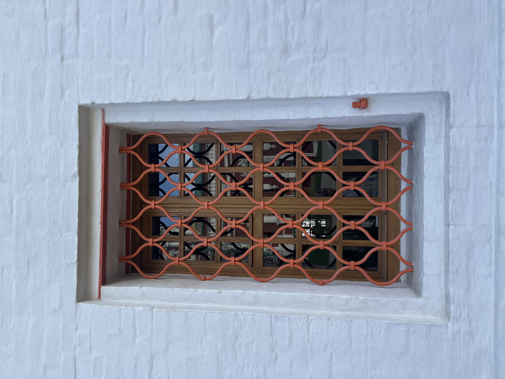 A close-up, slightly tilted shot captures a window with a distinctive decorative grate, set within a textured white wall. The window is recessed into the wall, framed by a reddish-orange border.
The wall itself is a rough, uneven white, suggesting a traditional plaster or whitewash finish over possibly brickwork. Covering the window is a prominent, reddish-orange metal grate, featuring an elaborate, repeating pattern of interconnected wavy or S-shaped loops, which serves both a protective and aesthetic function.
Behind the vibrant grate, a wooden window frame is visible, segmented into multiple smaller, rectangular glass panes. These panes reflect the surrounding environment, showing distorted views of what appear to be other architectural structures, possibly with red elements, and darker, undefined forms, perhaps trees or shadowed parts of buildings.
In the upper right corner of the window's recess, a small, reddish-orange, mushroom-shaped fixture is attached to the wall. The lighting is soft and diffused, indicating daytime with no harsh shadows, suggesting an overcast day or indirect light. The overall scene evokes a sense of historical architecture, consistent with a setting in Moscow, Russia. There is no visible text in the image.