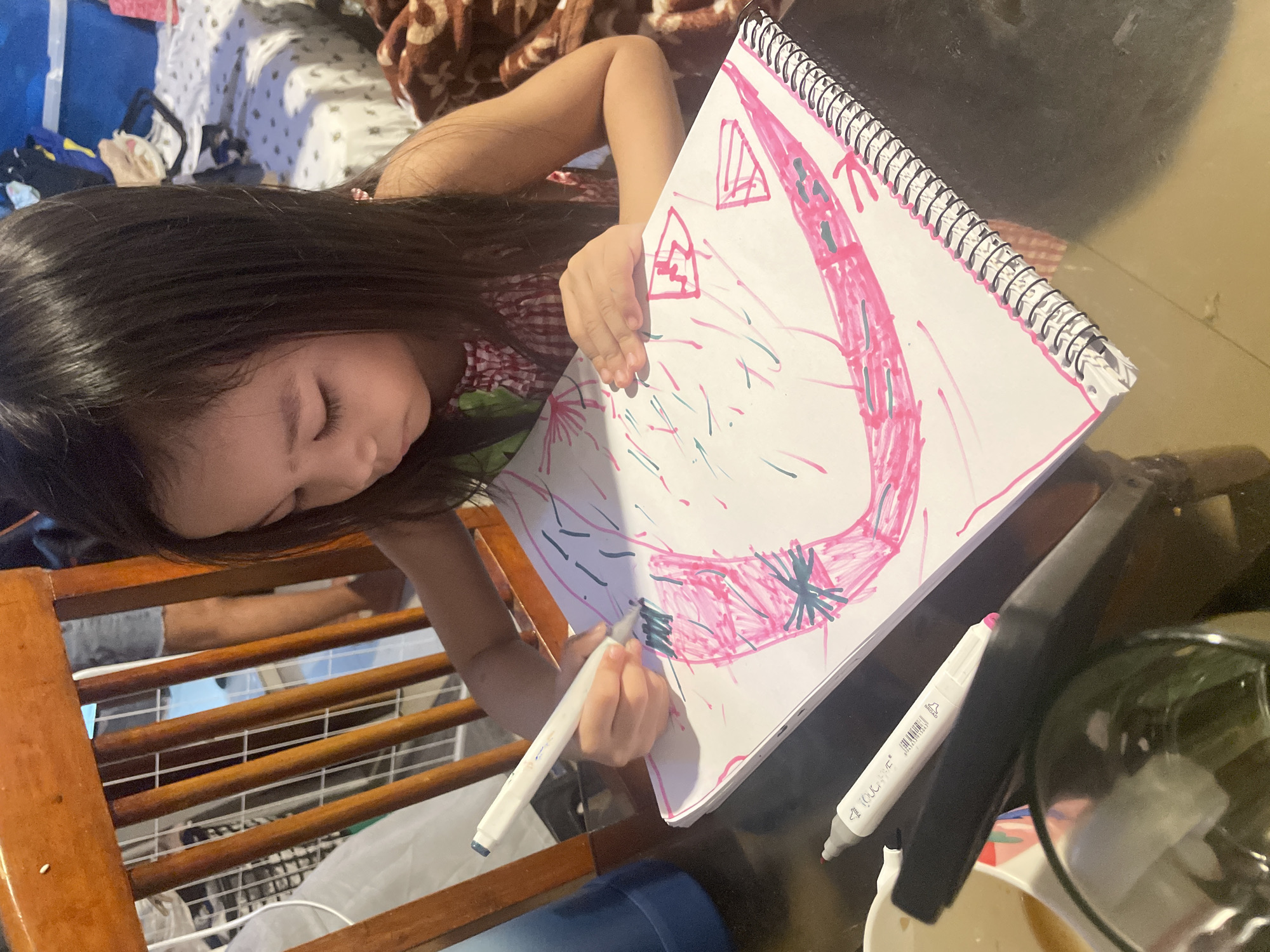 A young girl with long, dark hair is drawing on a white spiral-bound notebook with colorful markers. She is wearing a pink and white checkered dress. She is sitting at a table, and there are several markers on the table. The background shows a wooden chair and a bed with a patterned blanket. The lighting is natural, coming from a window or overhead light. The image appears to be taken indoors in a residential setting. There is no indication of the specific location within Davao City, Philippines.
