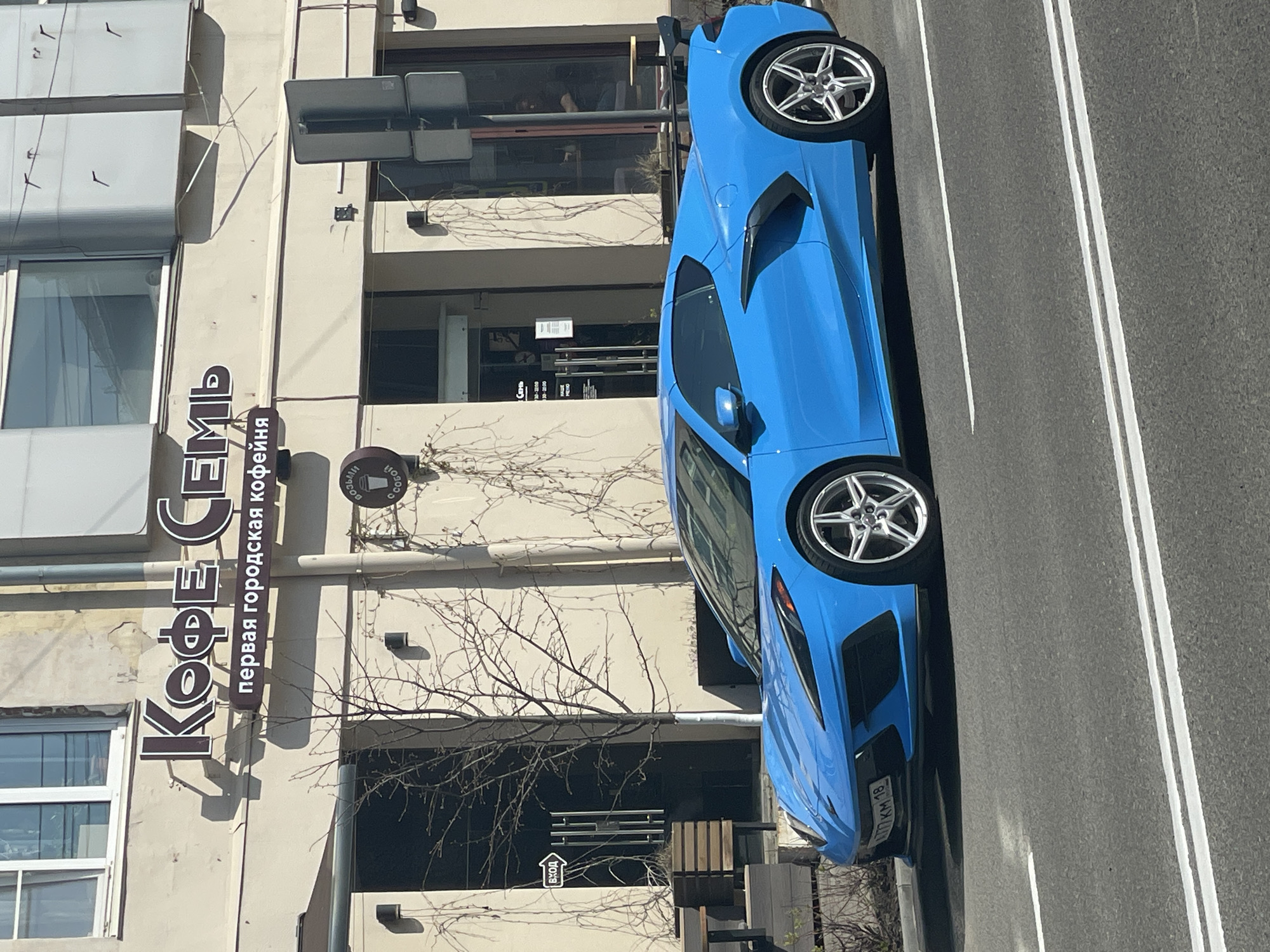 A striking bright blue sports car, likely a Chevrolet Corvette, is parked parallel to the curb on the right side of a street. The car faces away from the viewer. Two parallel white lines on the asphalt mark the street lanes.
To the left of the car is a multi-story building with a cream-colored facade. The building houses a coffee shop named "Кофе Семь" (Coffee Seven), as indicated by a vertical sign in Russian. The text below the coffee shop name reads "первая городская кофейня," meaning "first city coffee shop." The sign also includes the phrase "ВОЗЬМИ С СОБОЙ" (take with you). A small graphic with a coffee cup and steam adds to the signage. Bare tree branches are visible between the building and the car. The license plate of the car reads "777 KM 18".
The license plate "777 KM 18" indicates the car is registered in the Udmurt Republic, with the '18' denoting the region code. Given the Russian language sign, it is safe to assume this photograph was taken in Russia. The coffee shop name and information on the sign are clues that point to the location being in Izhevsk, the capital city of the Udmurt Republic.