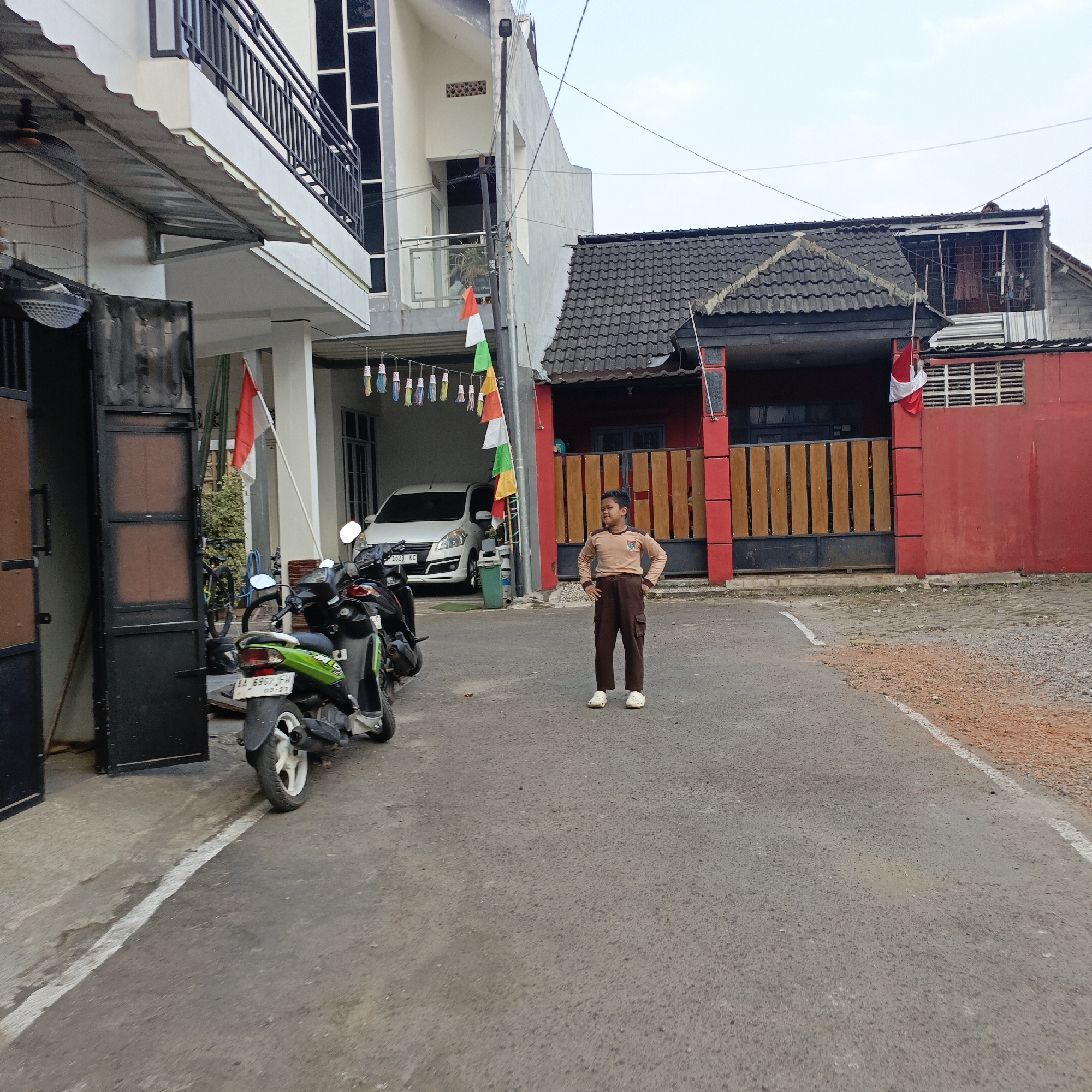 The image depicts a residential street scene in Salatiga, Indonesia, likely on a bright, overcast day. In the foreground, a young boy stands in the middle of the asphalt road, facing the camera with his hands on his hips. He is wearing a brown long-sleeved shirt with a crest on the chest and brown trousers, along with white slip-on shoes.
To the left of the boy, parked on the side of the road, are several motorcycles. A green and black scooter is most prominent, with its license plate visible. Behind it, other motorcycles are partially obscured, as well as a bicycle. Adjacent to the motorcycles, a large black gate is open, revealing a walkway and a white car parked inside a covered area. Above the walkway, an Indonesian flag is displayed on a white pole, and further along, strings of small, colorful decorative flags are hung.
To the right of the boy, a red house with a dark-tiled roof is visible. A wooden fence with vertical slats separates the property from the street. Another Indonesian flag is attached to the wall of this house. The building on the far left, behind the motorcycles and gate, is a multi-story structure with white walls and dark-framed windows, featuring a balcony with a black railing. Overhead, electrical wires crisscross the sky. The road surface is asphalt, with some gravel and dirt visible along the edges. The overall impression is of a typical urban or suburban residential area.