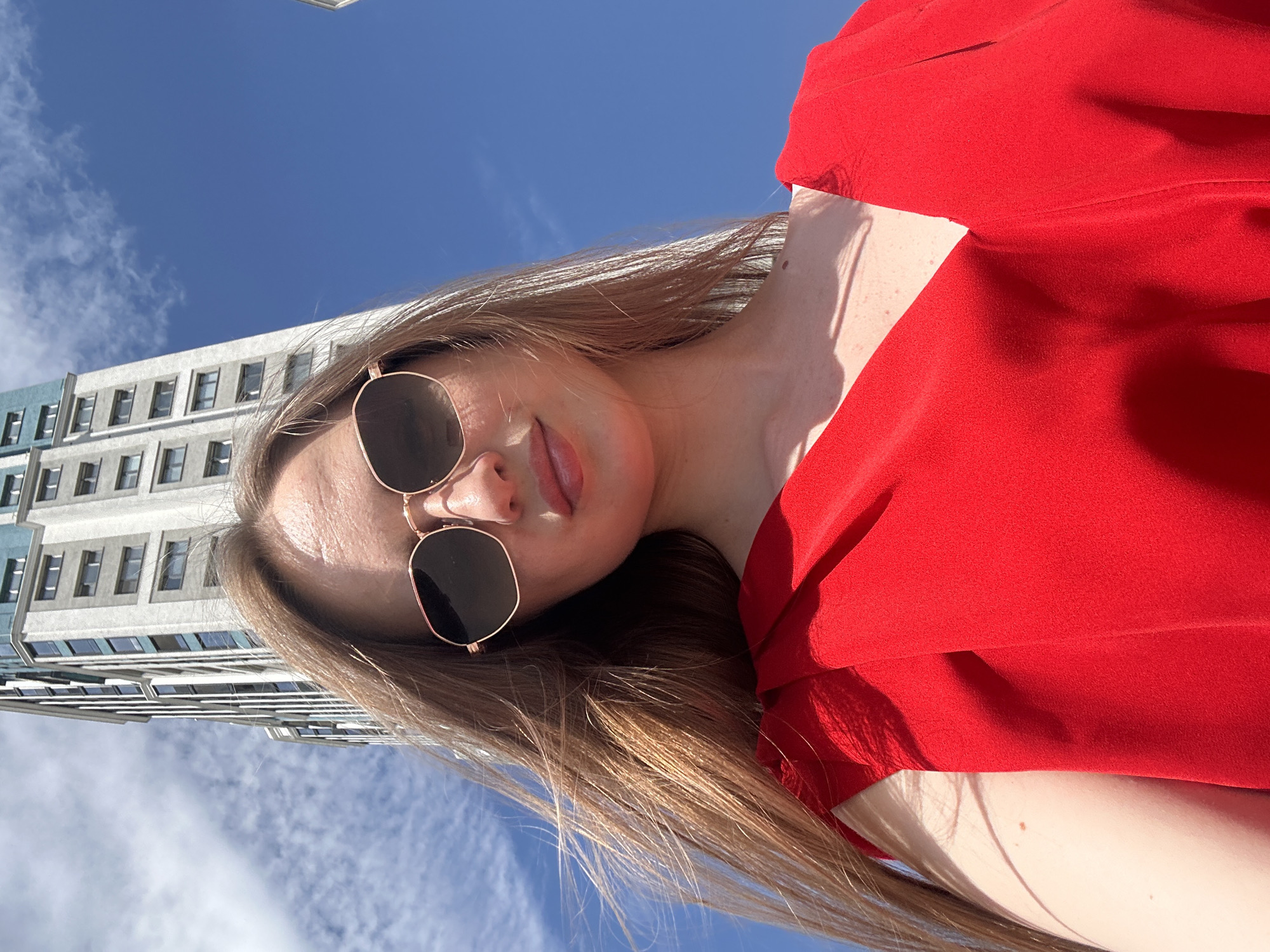 An outdoor, low-angle, medium shot features a fair-skinned woman with long, light brown hair and sunglasses. She is wearing a red v-neck dress and is looking directly at the camera. Behind her, a tall, modern, grey building with blue accents and numerous windows rises against a bright blue sky with scattered clouds. The woman's face is in focus, while the building is slightly blurred.