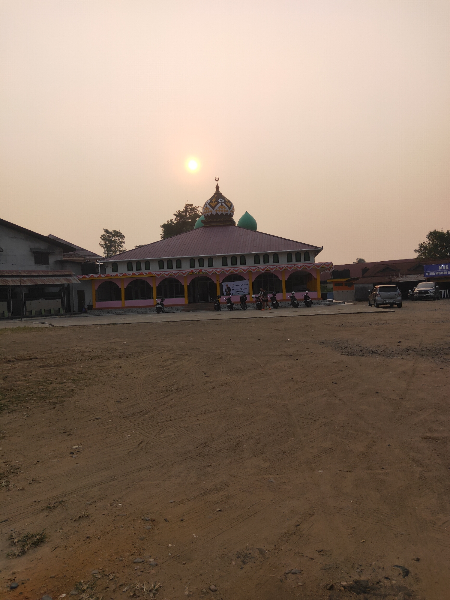 This is a photograph of the exterior of a mosque located in Sintang, Indonesia, during sunset. The sky is a hazy orange, and the sun is visible low on the horizon. The mosque features a red tiled roof with decorative yellow and pink arches along its facade. Two green domes and a smaller, more ornate dome are visible on the roof. Several motorcycles are parked in front of the mosque, and two cars are parked in the background. The foreground of the image is a dirt lot with tire tracks. A banner is visible on the side of the mosque.