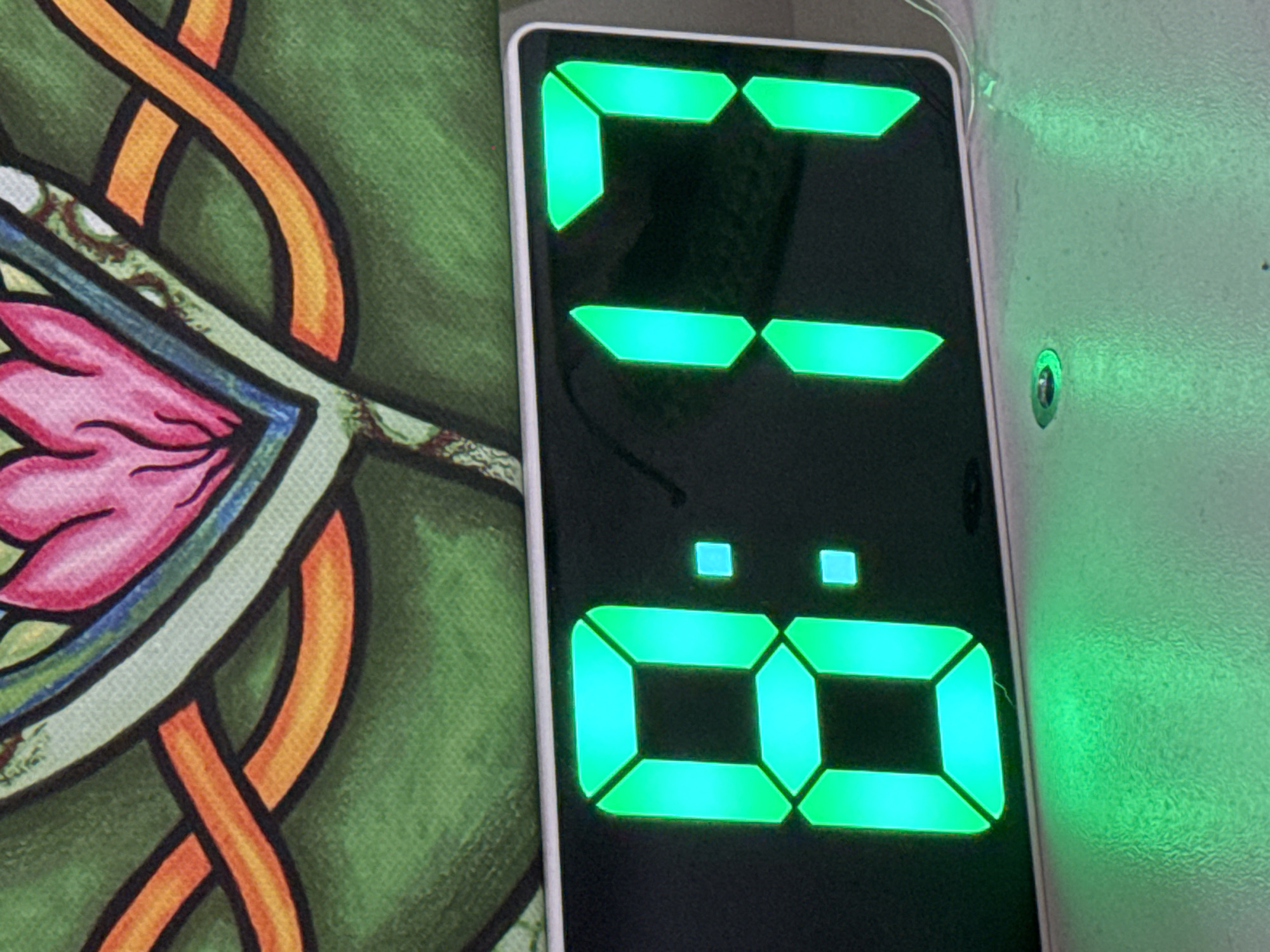 The image depicts a digital clock displaying the time 13:08. The clock has a black background with green LED digits. To the left of the clock is a portion of a patterned fabric with a predominantly green background, featuring orange and pink design elements. To the right of the clock is a plain, light-colored surface with a slight texture. The environment appears indoors with ambient lighting. There is no indication of location in Ketoshi, Russia or any other city.