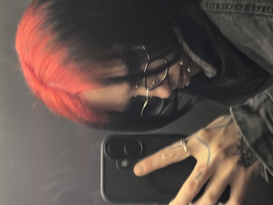 An individual is depicted in a mirror selfie taken within Plovdiv, Bulgaria. The subject's reflection shows a person with two-toned hair, featuring a vibrant red top transitioning to dark black. They wear thin-framed eyeglasses and multiple spike-like piercings around the lower facial area. A dark-colored jacket or similar outer garment is visible, along with a dark scarf or high collar. The subject's left hand holds a black smartphone, which has a dual-lens camera visible on its rear. A silver-toned ring is present on the ring finger of the visible hand, which also displays several tattoos, including linear designs and a darker pattern on the forearm. The background consists of an indistinct, dark, reflective surface, suggesting an indoor setting.