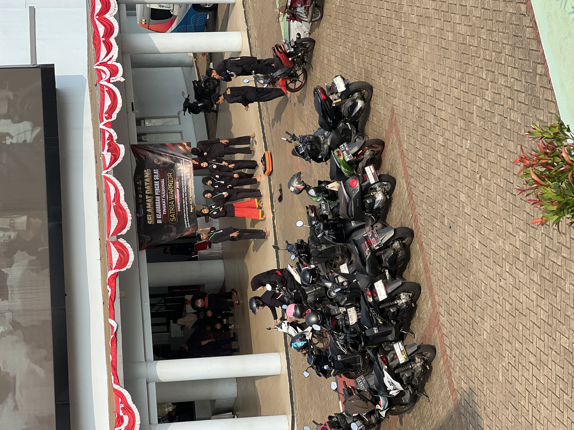 The image depicts an outdoor scene in Cibinong, Indonesia, likely at a venue hosting a Pencak Silat championship, as indicated by the banner that reads "SELAMAT DATANG DI KEJUARAAN PENCAK SILAT TINGKAT NASIONAL SATRIA WARRIOR CHAMPIONSHIP 2026". The setting is paved with interlocking bricks, with several parked motorcycles in the foreground. In the middle ground, a group of people in black uniforms, some wearing headscarves, stand in front of a building adorned with red and white decorative bunting. Some are interacting, while others appear to be waiting. Two individuals are seated on a motorcycle. A red and white vehicle, possibly an ambulance or support vehicle, is visible on the right side of the frame. The background shows a large screen or display mounted high on a building. The lighting suggests it is daytime with direct sunlight.