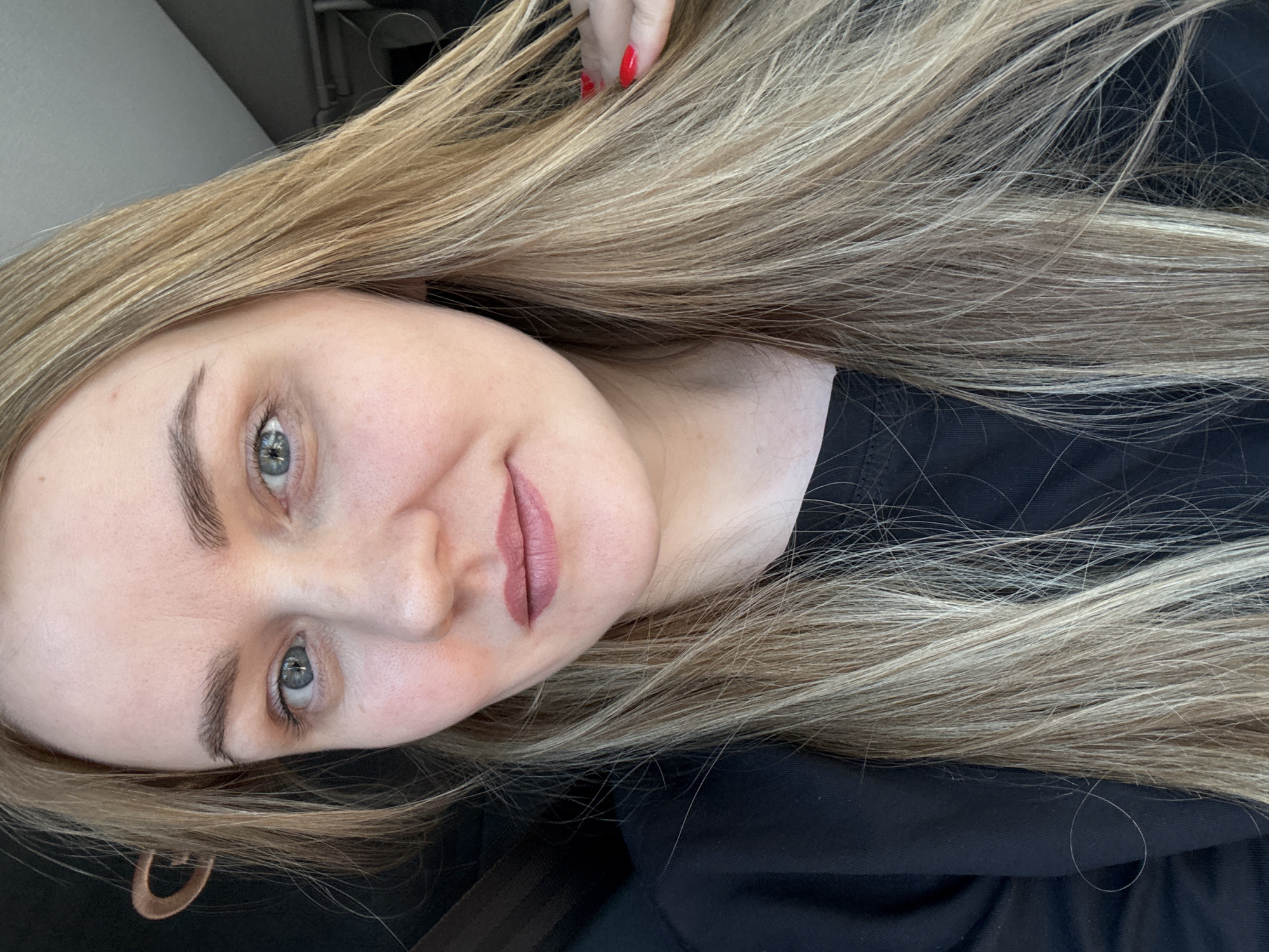 This image depicts a single female subject positioned for a frontal portrait, with the scene taking place in Iiul'skoe, Russia. The individual, appearing to be an adult, has long, straight blonde-to-light-brown hair that extends past her shoulders and covers parts of her black, long-sleeved top. Her skin is fair, and she has blue eyes. Subtle pink lipstick is applied to her lips, and her eyebrows are well-defined. Her gaze is directed towards the viewer, accompanied by a slight, closed-lip smile. Her right hand, adorned with red nail polish, is visible on the right side of the frame, touching or running through her hair. The background consists of a dark, indeterminate object on the left, possibly a car seatbelt or interior panel, and a lighter, muted grey area on the right, suggesting an indoor or vehicle environment with ambient lighting.