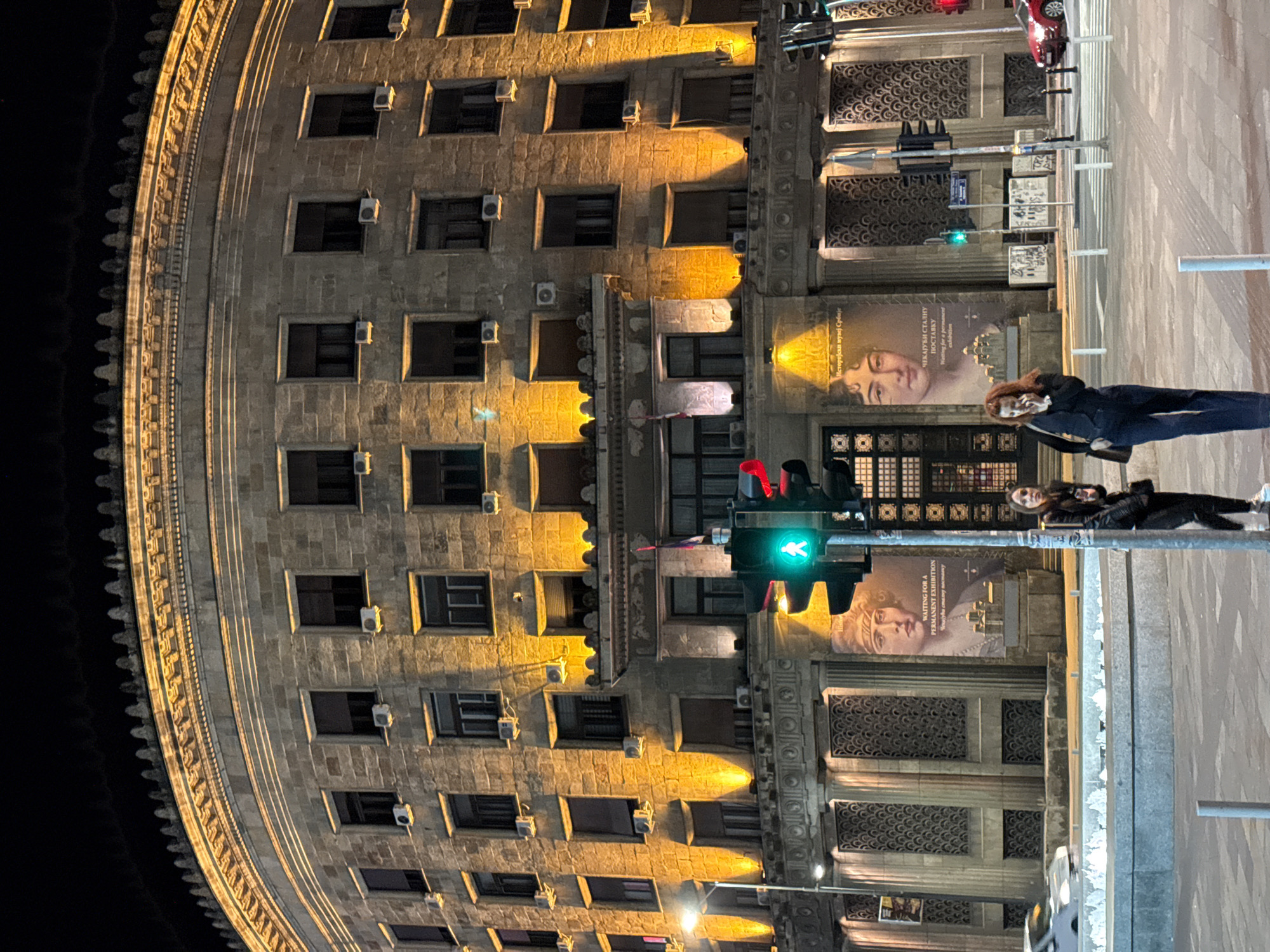 The image shows a tall, illuminated building in Belgrade, Serbia, at night. The building, made of light-colored stone, has numerous windows and ornate detailing. Air conditioning units are visible on the exterior. A traffic light with a green walking symbol is illuminated in the foreground. Two people are standing on the sidewalk. One person is wearing a dark jacket and pants, while the other is in a black jacket and black pants. A large advertisement, possibly for the Historical Museum of Serbia, features a woman's portrait and the phrase "WAITING FOR A PERMANENT EXHIBITION" in both Serbian and English. The street appears wet, reflecting the ambient light.