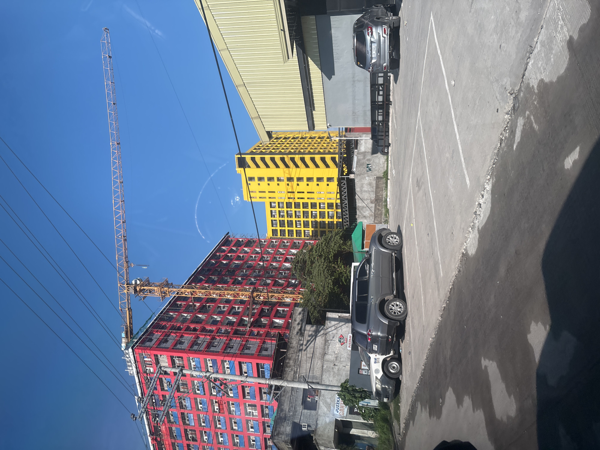 A sunny daytime, outdoor, wide-angle shot captures a street scene in Davao City, Philippines, dominated by construction and urban infrastructure. In the foreground, a concrete parking lot with white painted lines shows several parked vehicles. A dark grey SUV is parked facing away from the viewer in the right side of the frame. To its left, a lighter grey SUV is parked, with a white vehicle partially visible behind it. The ground in the parking lot appears wet in places, with some debris and cracks along the edges.
Behind the parking lot, a large, multi-story building with a yellow facade stands prominently. To the left of the yellow building, a taller, red-colored building with blue window frames is under construction, with a yellow tower crane extending over it. The red building is densely packed with balconies. Electrical wires crisscross the sky above the scene. A low-rise building with a grey facade is situated in front of the red building.
The background reveals more of the urban landscape, including what appears to be a warehouse-style building with a pale yellow corrugated metal roof. The sky is a clear, bright blue. The overall impression is one of ongoing development within a bustling city environment.