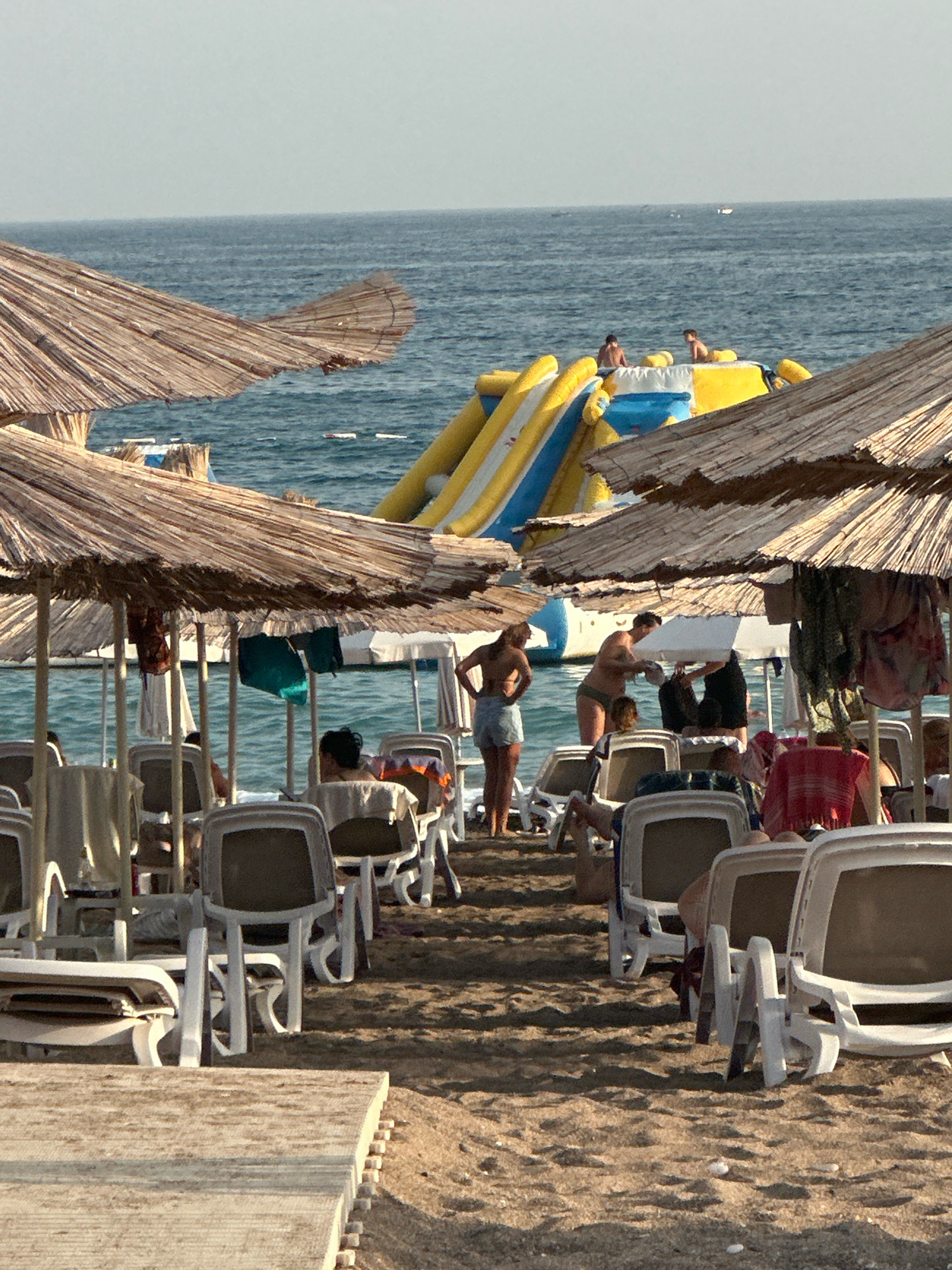 In Boreti, Montenegro, a crowded beach scene unfolds under a hazy sky. The foreground is dominated by the texture of sand and the weathered wood of a beach platform. Behind it, a row of empty white plastic lounge chairs, some adorned with towels, are neatly arranged under thatched umbrellas.
Further back, more lounge chairs and umbrellas are visible, some occupied by people. To the right, a group of individuals are gathered near the water's edge. One person, wearing a green bikini bottom and a dark top, is tending to another person partially obscured by a white umbrella. To their left, a person in denim shorts and a dark top stands with their hands on their hips, facing the water.
In the background, the blue ocean stretches towards the horizon, dotted with a few small white buoys. A large inflatable water slide, yellow and blue, is positioned in the water, with two figures visible near its top. The overall impression is one of a bustling beach resort on a sunny day.