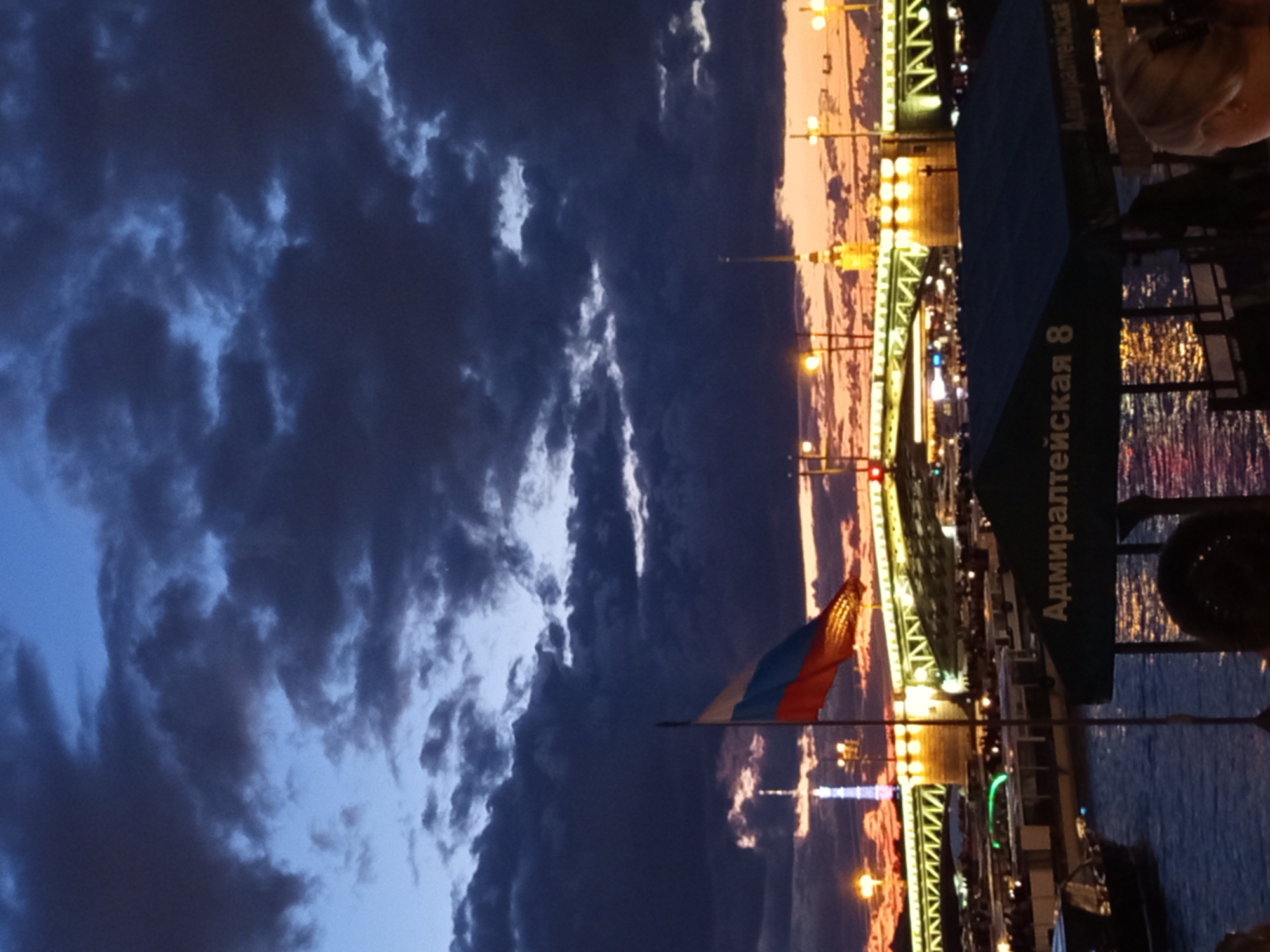 A Russian flag flies from a pole in the foreground, positioned in front of a wide, illuminated bridge spanning a body of water. The sky above is dark and cloudy, with a dramatic sunset visible behind the bridge, casting orange and yellow hues. Several boats are docked or moving on the water, some appearing to be passenger vessels. A building with a tall spire is visible in the distance, beyond the bridge. A dark green awning with white lettering that reads "Адмиралтейская 8" is partially visible in the lower right portion of the image, obscuring a section of the boats and water. The scene appears to be a cityscape at dusk, likely in a location with significant water traffic and architectural landmarks.