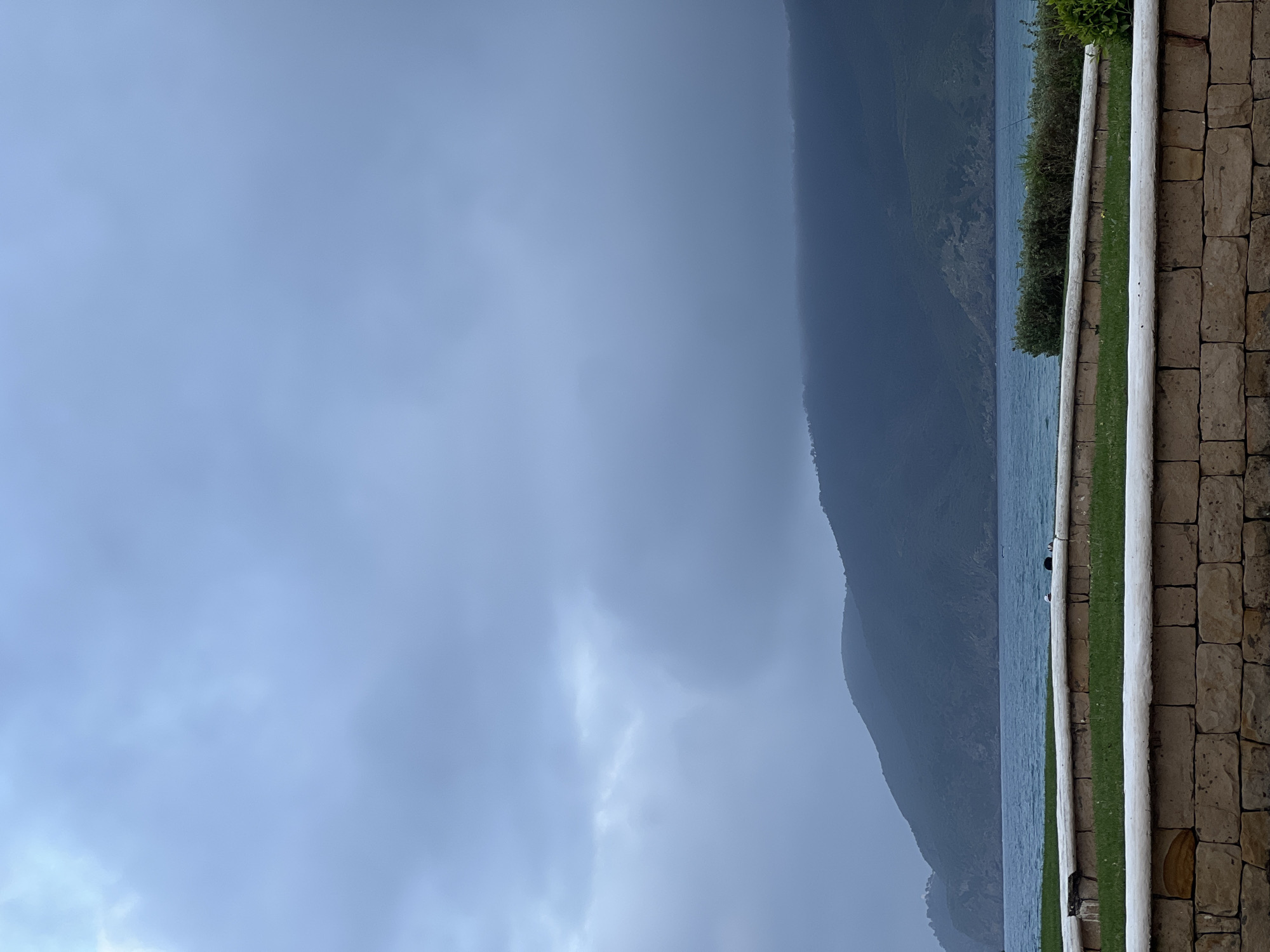 The image shows a cloudy, overcast view of what appears to be a mountainous coastline with water meeting the land. The location is likely in or near M'Diq, Morocco given the request. The foreground exhibits a wall constructed of light-colored stone blocks with white poles running vertically along its edge, and a strip of green grass runs behind the wall. In the midground, there is water which meets the shore, followed by the dark silhouette of what appears to be a coastal mountain or hill. The sky is overcast and heavily clouded. No people are visible in the image.