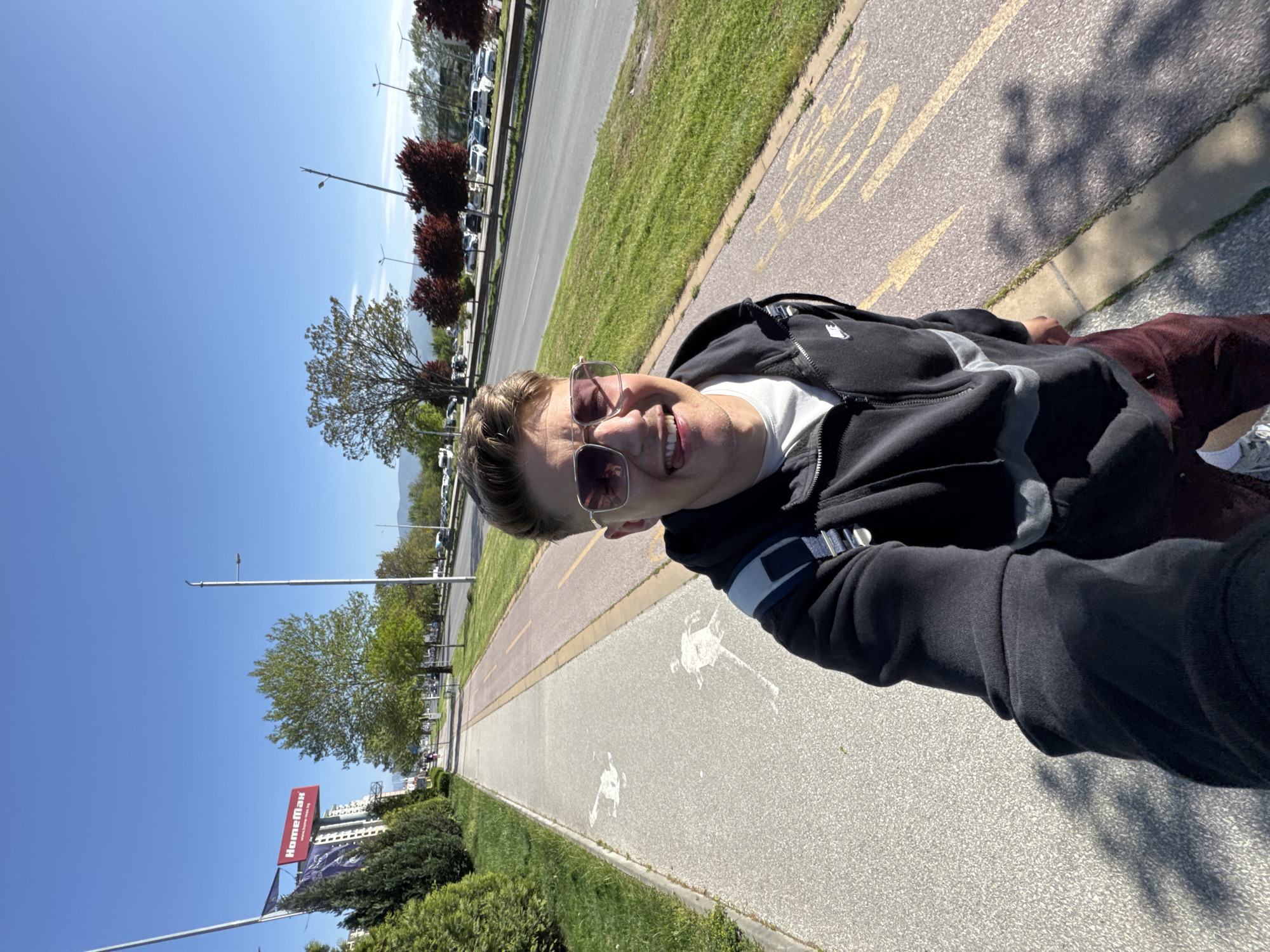 A young man is taking a selfie on a sunny day in Plovdiv, Bulgaria. He is on a paved path, next to a grassy area and a road with parked cars. A building with a "HomeMax" sign is visible in the background. The path has markings indicating it is a bicycle lane. The man is wearing a dark-colored jacket and sunglasses. He appears to be smiling.