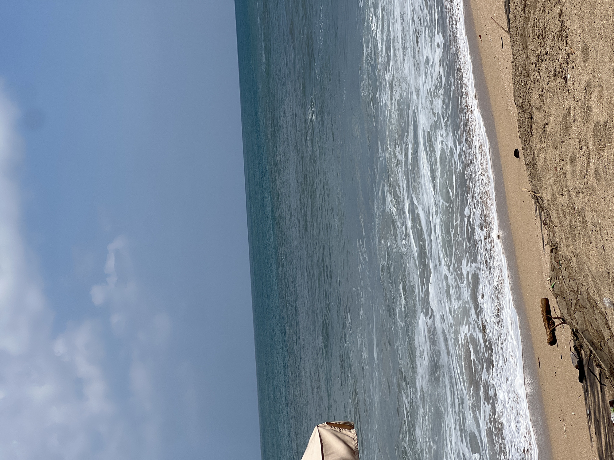 The image captures a tranquil scene on the beach in Kuta, Indonesia. The vast expanse of the ocean, with its gentle waves and the horizon meeting the sky, dominates the upper portion of the frame. The water exhibits a serene blue-green hue, contrasting with the lighter, foamy surf that laps at the shore. A small portion of a beige beach umbrella or canopy is visible on the far left, partially submerged in the water near the shoreline. The sandy beach occupies the foreground, with scattered debris, including pieces of wood and some dark objects, lying on the sand. The overall environment is one of natural beauty, with a clear, bright sky and calm sea.