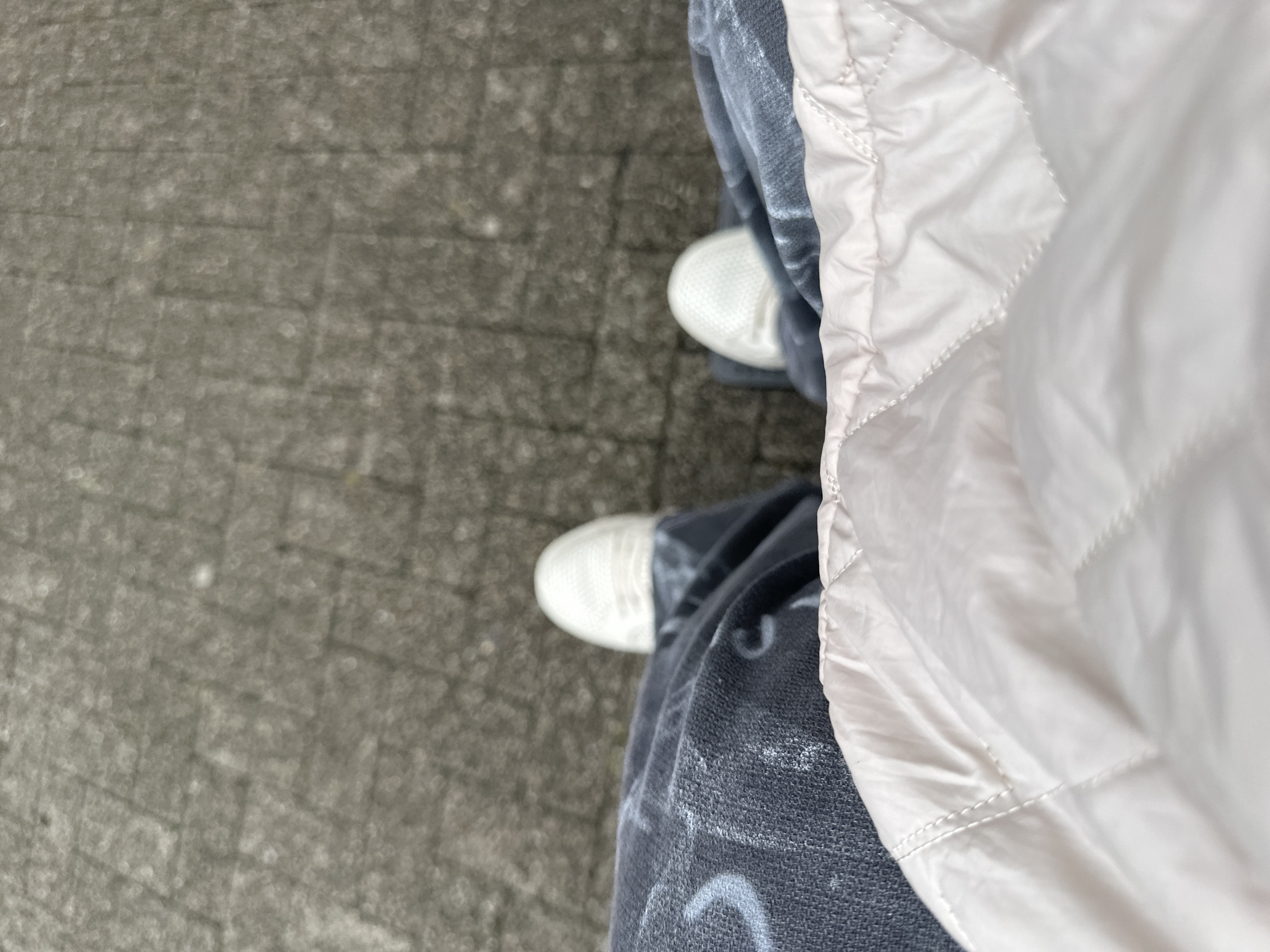 A first-person perspective shot of a person standing on a paved walkway. The walkway is made of grey rectangular pavers arranged in a grid pattern. The person is wearing dark wash jeans with a white graphic print on the left leg. White sneakers are visible on both feet, with the left shoe more prominently displayed. The lower portion of the frame is filled with a light pink, quilted puffer jacket or vest. The scene appears to be outdoors in an urban environment, possibly Krefeld, Germany, given the context. No other individuals or specific landmarks are visible.