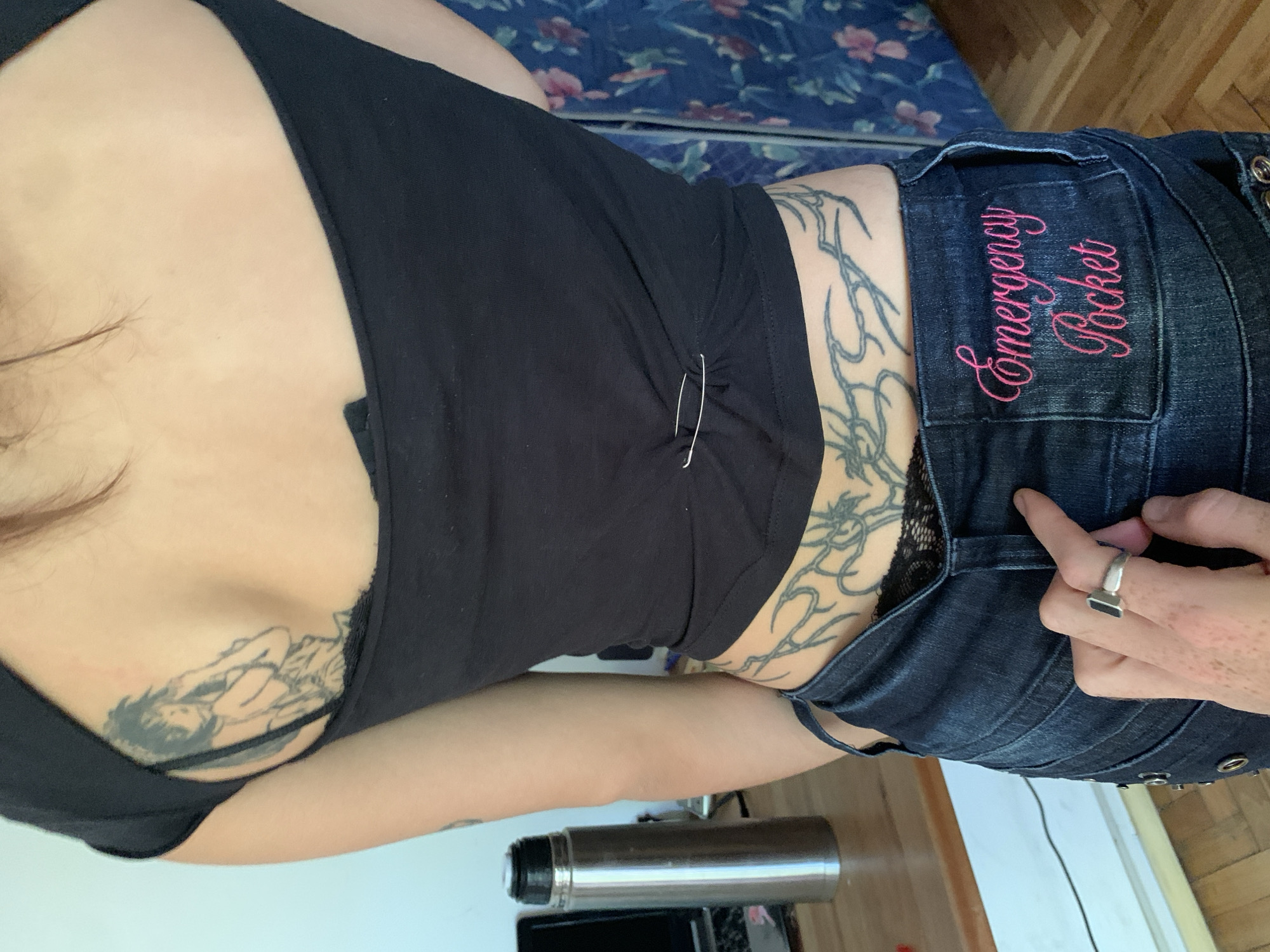 The image displays a partial view of a person, likely female-presenting, lying down indoors, possibly in a bedroom in Buenos Aires, Argentina. The perspective is from above, looking down the length of the body. The individual is wearing a black tank top or camisole, cinched at the side with two silver safety pins, and dark blue denim jeans. A strap of black lace underwear is visible under the top on the right side.
Several tattoos are prominent: a large, intricate tribal-style design covers the person's right side and lower back, extending towards the hip. Another tattoo on the left chest depicts an anime or manga character in a reclining pose, possibly sleeping. The person's right hand, wearing a silver ring with a black rectangular stone, rests near the back right pocket of their jeans. This pocket is embroidered with the words "Emergency Pocket" in pink script.
The background includes a bed with a blue sheet featuring a floral pattern and a wooden floor. A silver thermos or bottle and a small black electronic device are visible on the floor. Dark hair is visible on the left side of the frame, belonging to the person. The lighting suggests it is daytime.