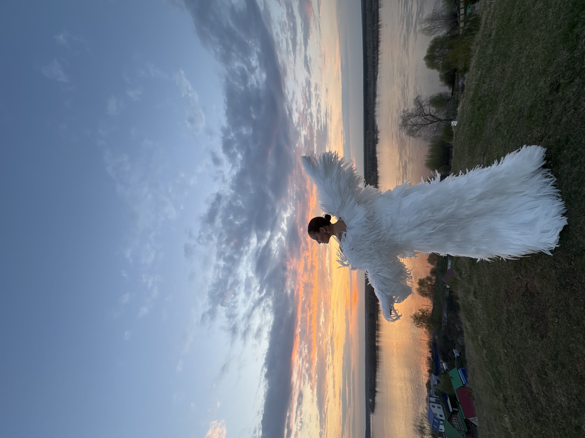 A woman stands on a grassy field near a body of water, possibly the Ilovlya River, wearing a white feathery garment that covers her entire body, except for her head, which is positioned in profile. The setting sun casts a warm orange glow on the water and the nearby landscape. In the background, there is a cityscape, likely Olkhovka, with some houses visible. The sky is a mix of blue and grey clouds.