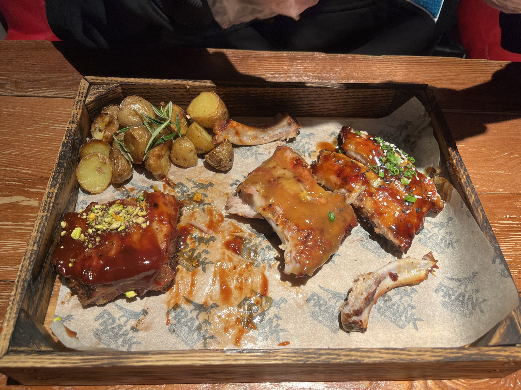 A wooden serving tray, lined with grease-stained parchment paper featuring faint graphic patterns, displays a partially consumed meal. The tray contains several pieces of cooked ribs coated in various sauces; one prominent piece is covered in a dark red sauce topped with green crushed pistachios, while other smaller pieces are sauced and garnished with sesame seeds and chopped green onions. A cleaned rib bone is present, indicative of consumption. Alongside the ribs are multiple small, roasted potatoes, some whole and some halved, garnished with fresh rosemary sprigs. The tray rests on a dark brown wooden surface, possibly a table, with distinct shadows suggesting an overhead light source. To the immediate right of the tray, a dark fabric object and a red object are partially visible. This scene is located in Родниковское сельское поселение, Ukraine.