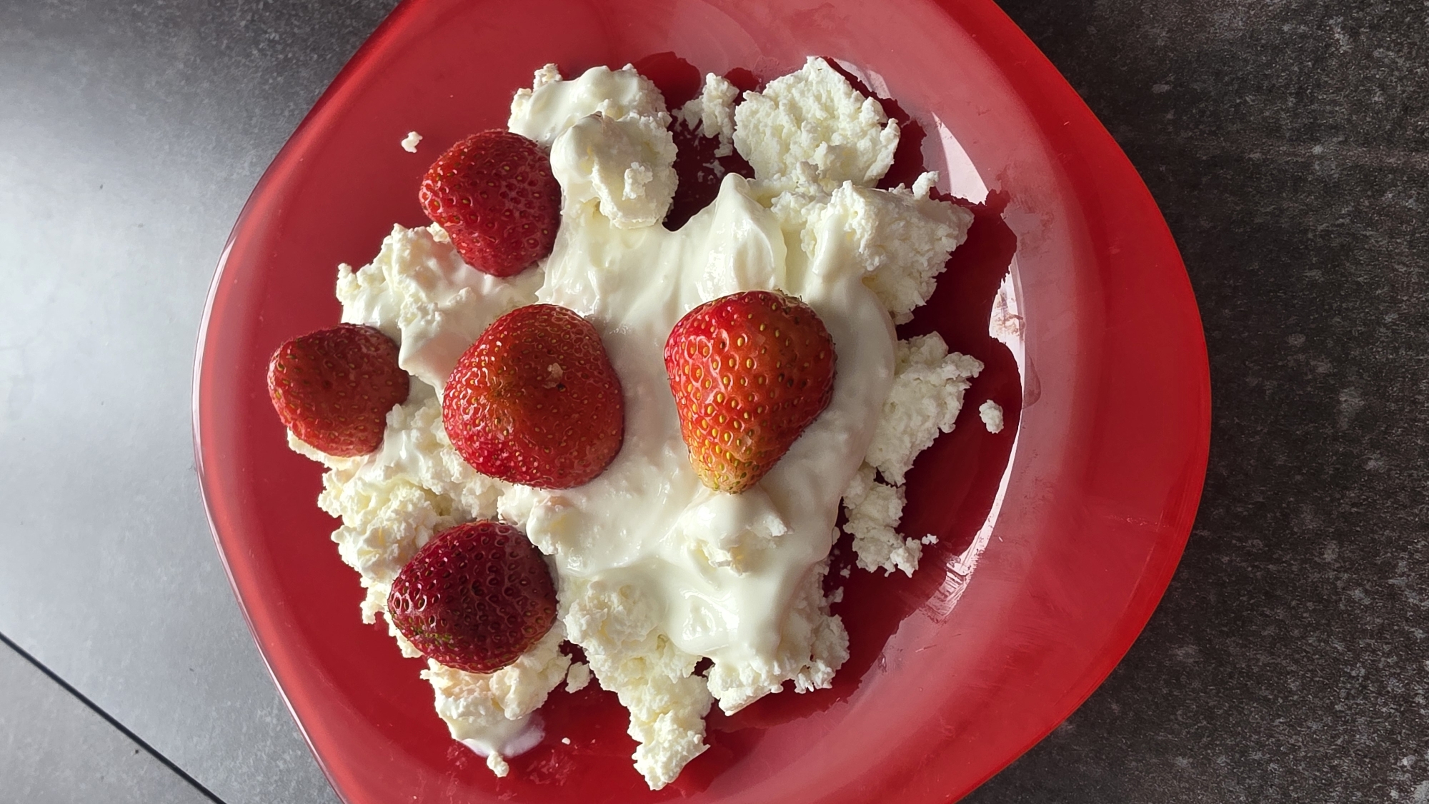 A close-up, high-angle shot shows a red plate filled with cottage cheese and several strawberries. The cottage cheese is piled in a heap in the center of the plate, with a swirl of creamy yogurt or sour cream on top. Five whole strawberries are scattered across the cottage cheese, some with their green tops still attached. The plate sits on a dark, speckled surface, possibly a countertop.