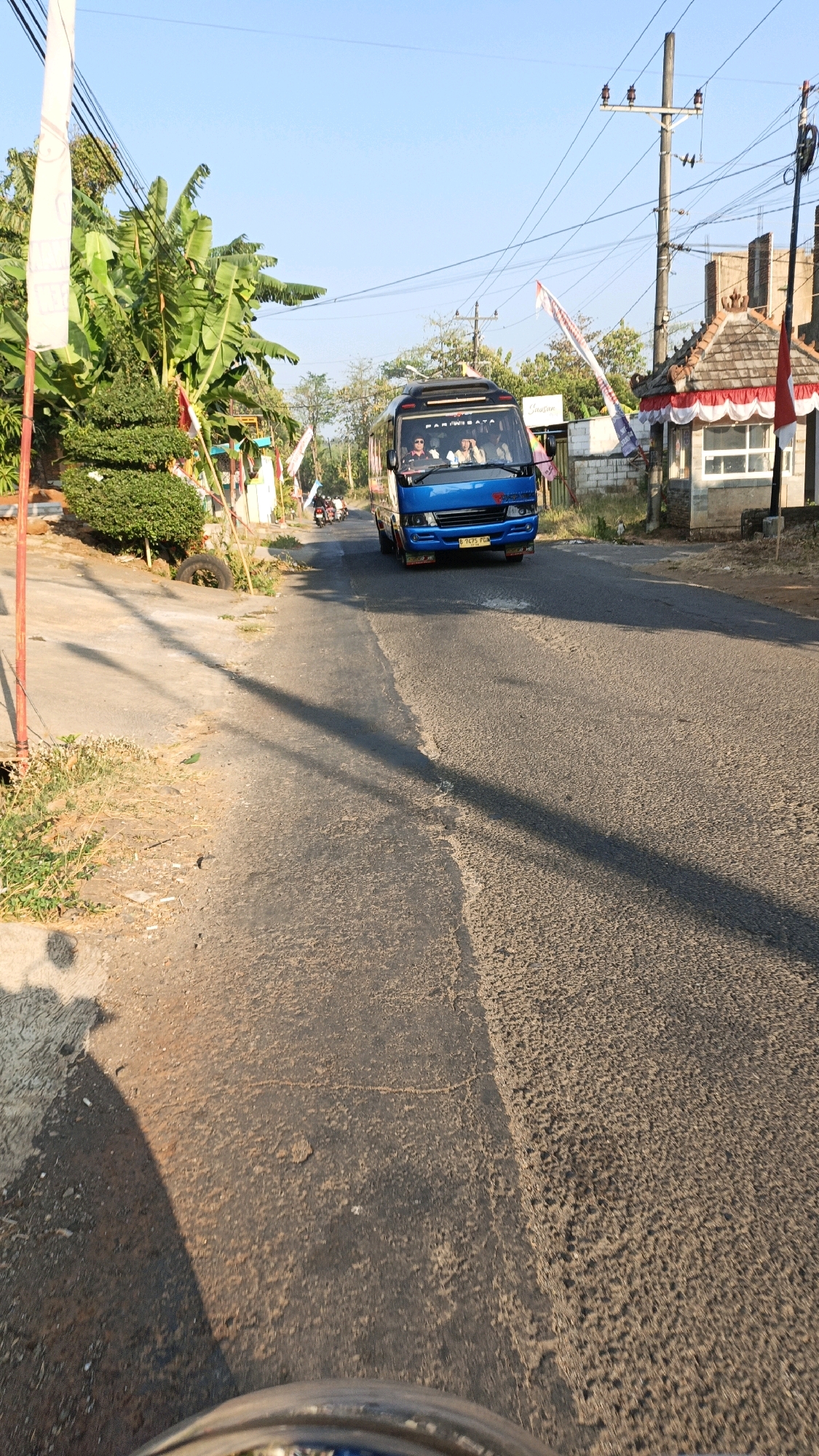 The image captures a bright, sunny day on a road in Jepara, Indonesia, likely a rural or suburban area given the vegetation and the presence of a small roadside structure. A blue minibus, possibly a tour bus or public transport, is the main subject, driving towards the viewer. The bus has several passengers visible inside, and the text "PARIWISATA" (tourism in Indonesian) is visible on its front.
On the left side of the road, there is a clear pathway with a red pole and some green bushes and banana trees. A large tire is visible near the edge of the pathway. Flags, likely Indonesian flags, are planted along the roadside and appear to be part of some local celebration or decoration.
To the right of the road, there is a small, ornate building with a tiled roof, possibly a ticket booth or a small shop, decorated with red and white fabric. Electric poles and wires are visible in the background, indicating the presence of infrastructure. The road surface is asphalt, with some patches of wear and tear. The overall environment suggests a warm climate. The photo is taken from a low angle, possibly from a moving vehicle, creating a dynamic perspective.