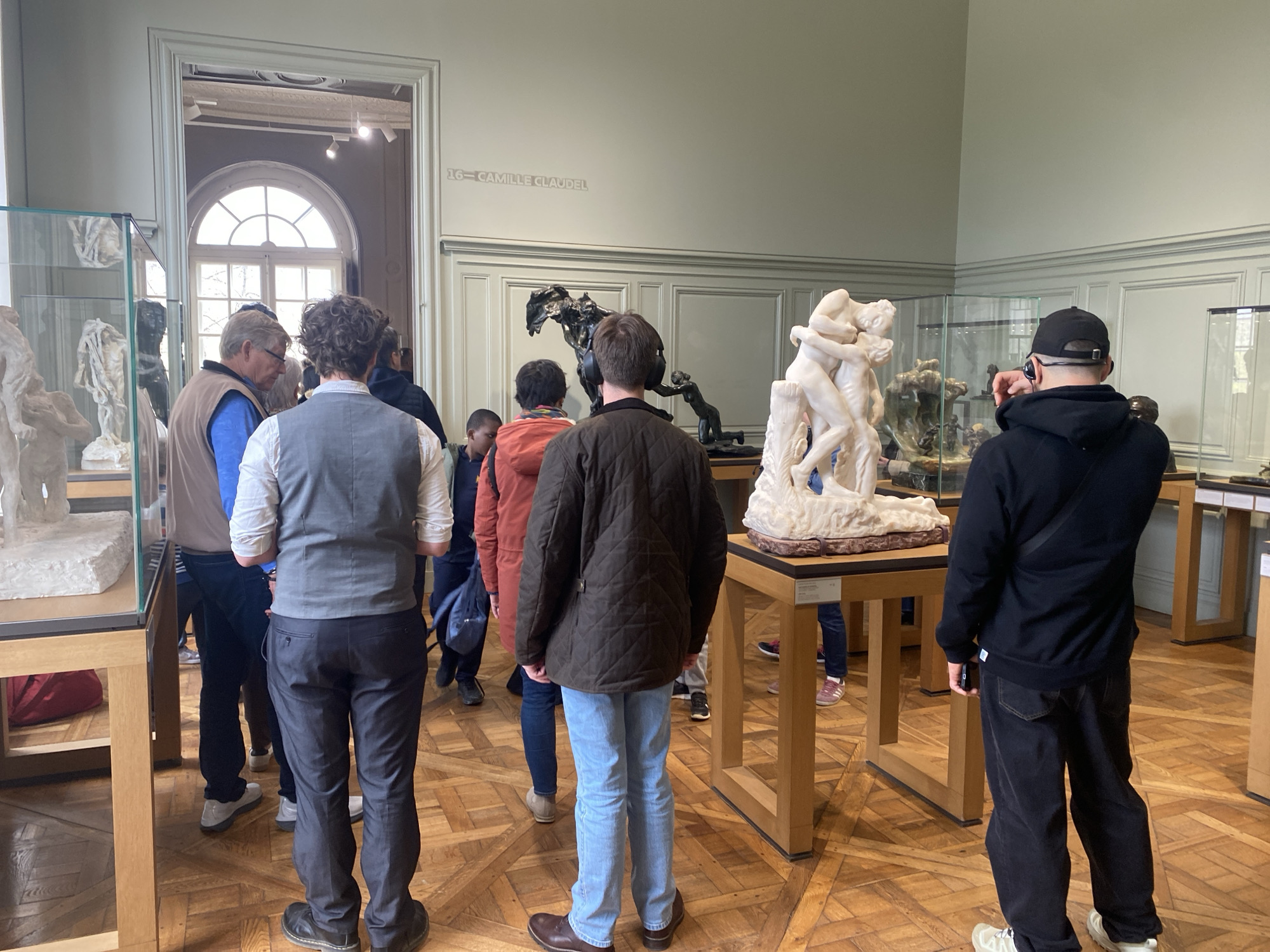 The scene depicts an indoor museum gallery, identified by wall text as "16 — CAMILLE CLAUDEL," suggesting the Musée Rodin in Paris, France. The room features light green walls with wainscoting, an ornate wooden parquet floor, and a large arched window in the background providing natural light. Approximately nine individuals are present, observing sculptures.
In the foreground right, a large white marble sculpture of two embracing figures stands on a light wooden pedestal. Behind it, several darker, patinated or bronze sculptures are displayed on similar pedestals, including a multi-figure piece and a smaller standing figure. On the left side of the room, additional sculptures are visible within glass display cases.
Multiple visitors are distributed throughout the space. In the foreground left, a man with curly brown hair wears a white shirt, grey vest, and dark trousers, facing away from the camera. To his right, an older man with light hair wears a light brown vest over a blue shirt, looking towards the left. A man in a dark brown quilted jacket and light blue jeans stands in the middle, facing the central sculptures. In the foreground right, a man wearing a black baseball cap, black hoodie, and dark pants holds a black smartphone in his right hand, pointed towards the artworks, suggesting he is taking a photograph. Other visitors, dressed in casual attire including an orange jacket, are partially visible in the mid-ground, also observing the exhibits. All individuals are focused on the artworks; no direct personal interactions are clearly depicted.