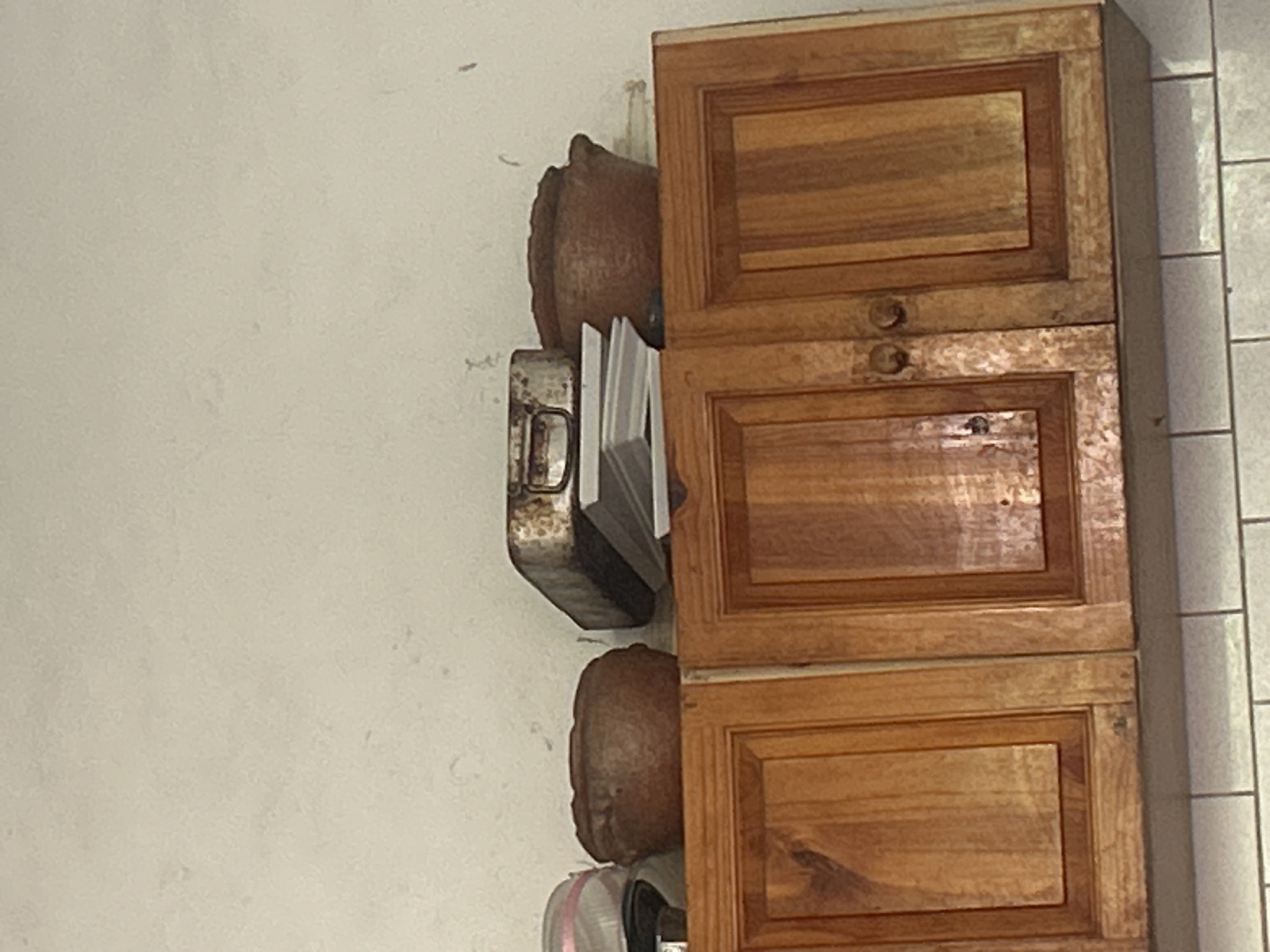 A low-angle medium shot captures an interior scene featuring a portion of a kitchen cabinet with items stored on top. The cabinet is made of wood and features two doors with visible grain. Above the cabinet, on a surface that appears to be the top of the cabinet or a shelf, are several objects. To the left is a glimpse of a round, brown ceramic pot. Next to it is a rectangular metal baking pan with a distressed, possibly burnt, surface. Stacked on top of the baking pan are several white plates. To the right of the baking pan is another round, brown ceramic pot with a similar texture to the one on the left. The wall above the objects is a neutral, off-white color with some minor imperfections. The scene is likely indoors, possibly in a residential kitchen. The lighting suggests ambient interior light, creating soft shadows. The image does not provide any specific landmarks or details that would allow for pinpointing a location within Santa Ana, Venezuela.
