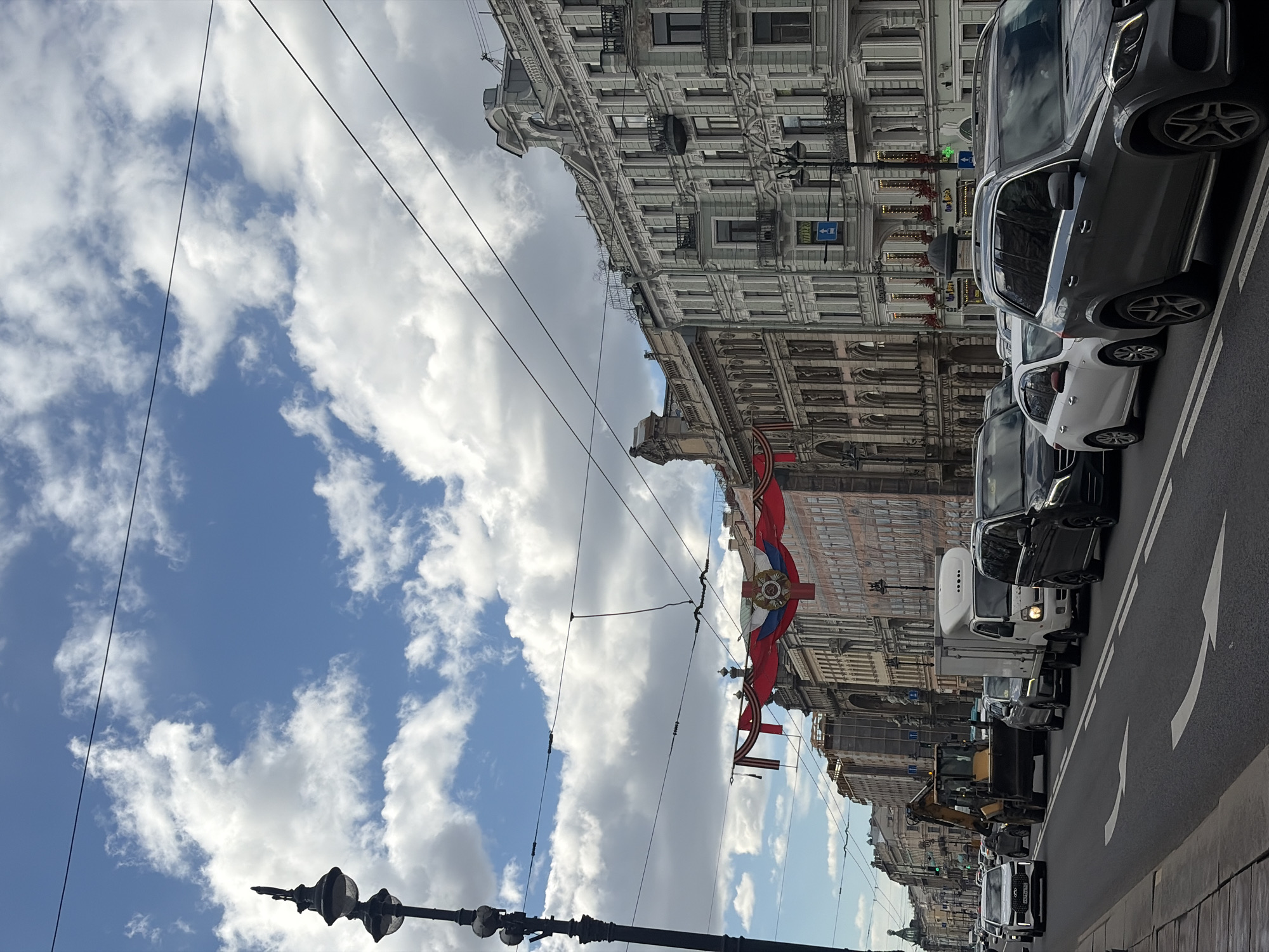 The image captures a street scene in Saint Petersburg, Russia, during what appears to be a festive or commemorative occasion. The sky is partially cloudy, with patches of blue visible. Buildings on the right side of the frame exhibit ornate, classical-style architecture. Russian flags are displayed prominently. Traffic lines the street. An excavator is among the vehicles.