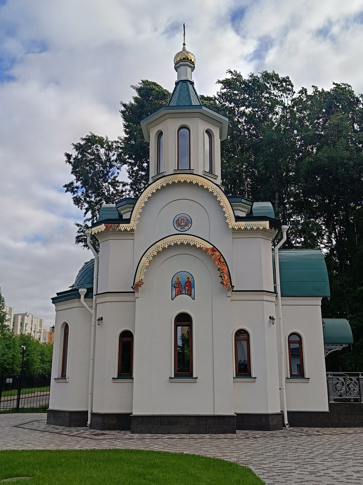 The image shows the Church of the Iberian Icon of the Mother of God, a relatively new Orthodox church located at 44 Vyborgskoye Highway in Saint Petersburg, Russia. The church is a modern, white building with a green roof and a golden dome. It features arched windows and decorative elements above the entrances. A manicured lawn and paved walkway are visible in the foreground, with a black metal fence to the left and trees to the right and behind the structure. In the background, residential buildings can be seen. The sky is cloudy. There are no people visible in the image.